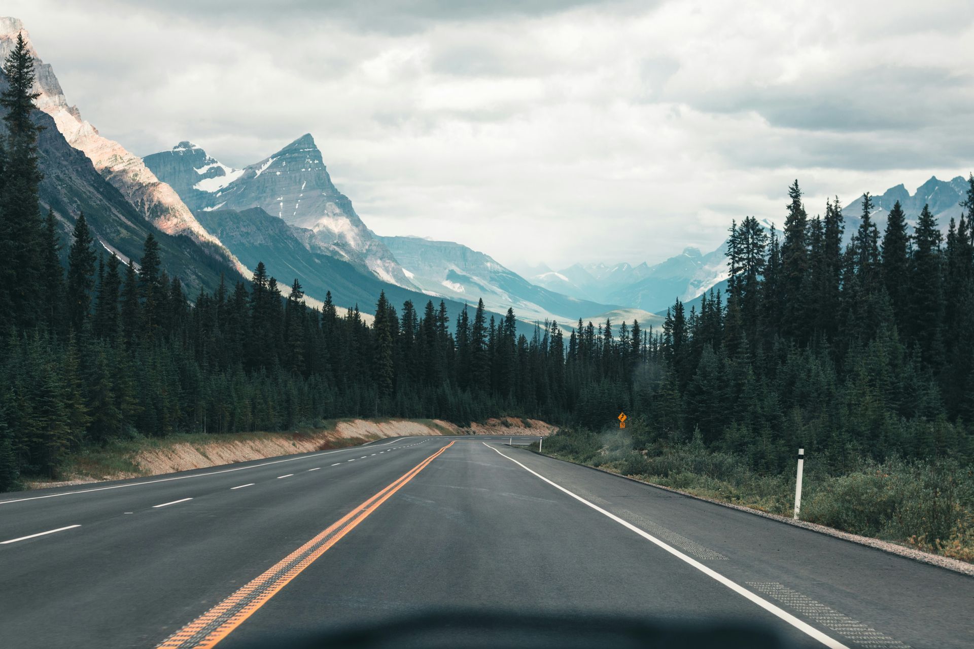 Road through a mountain pass with evergreen trees, snow-capped peaks, and a cloudy sky in Jasper, Canada.
