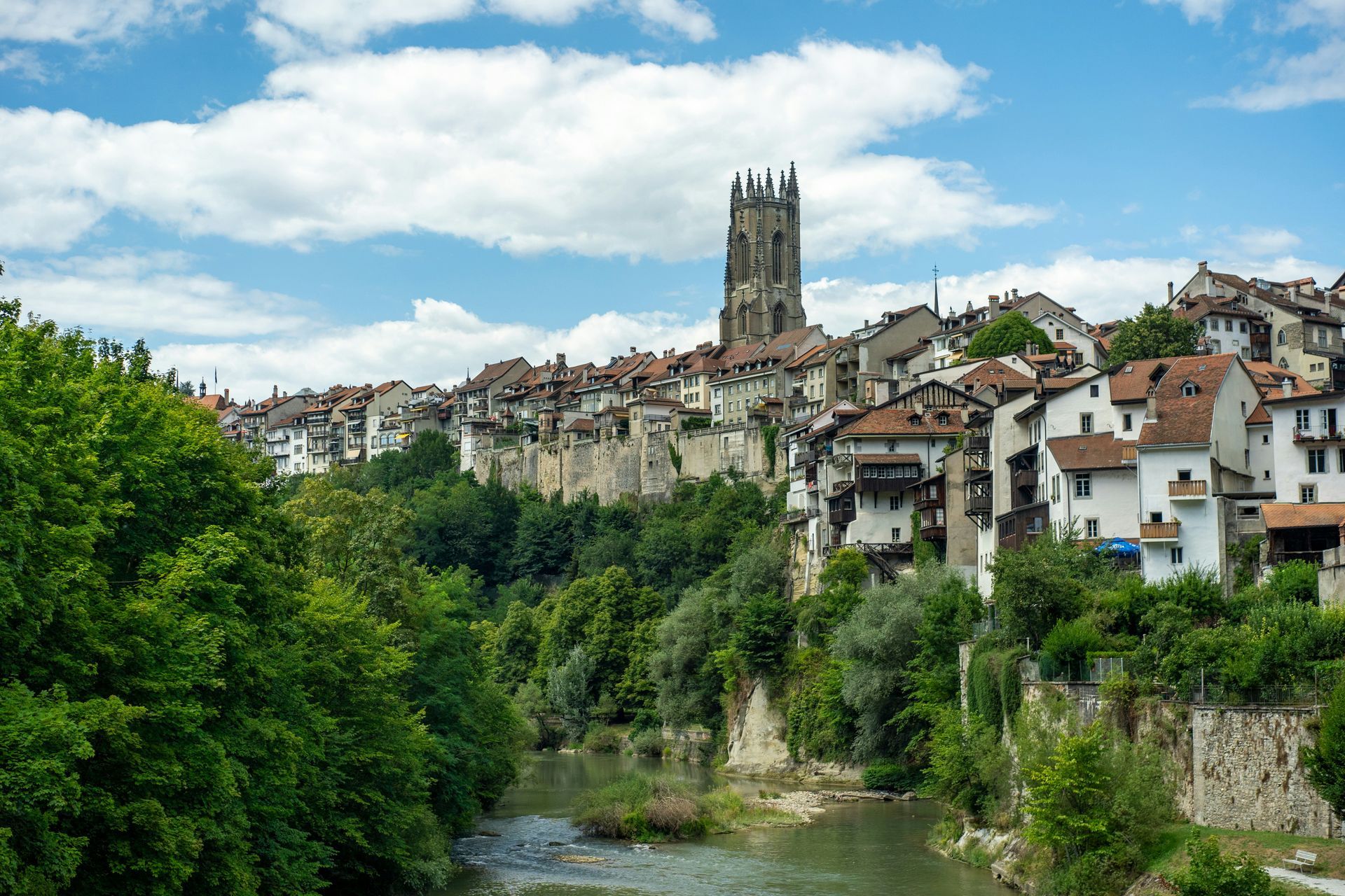 Cityscape of Fribourg, Switzerland, with a medieval church tower above white buildings and green trees, and a river.