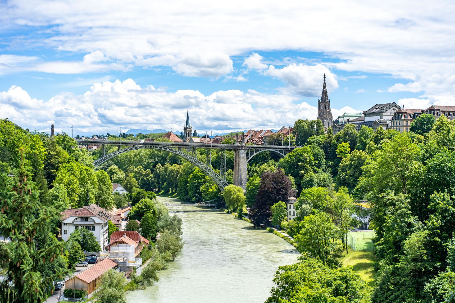 A scenic view of Bern, Switzerland, with a river, bridge, buildings, and green trees under a cloudy sky.