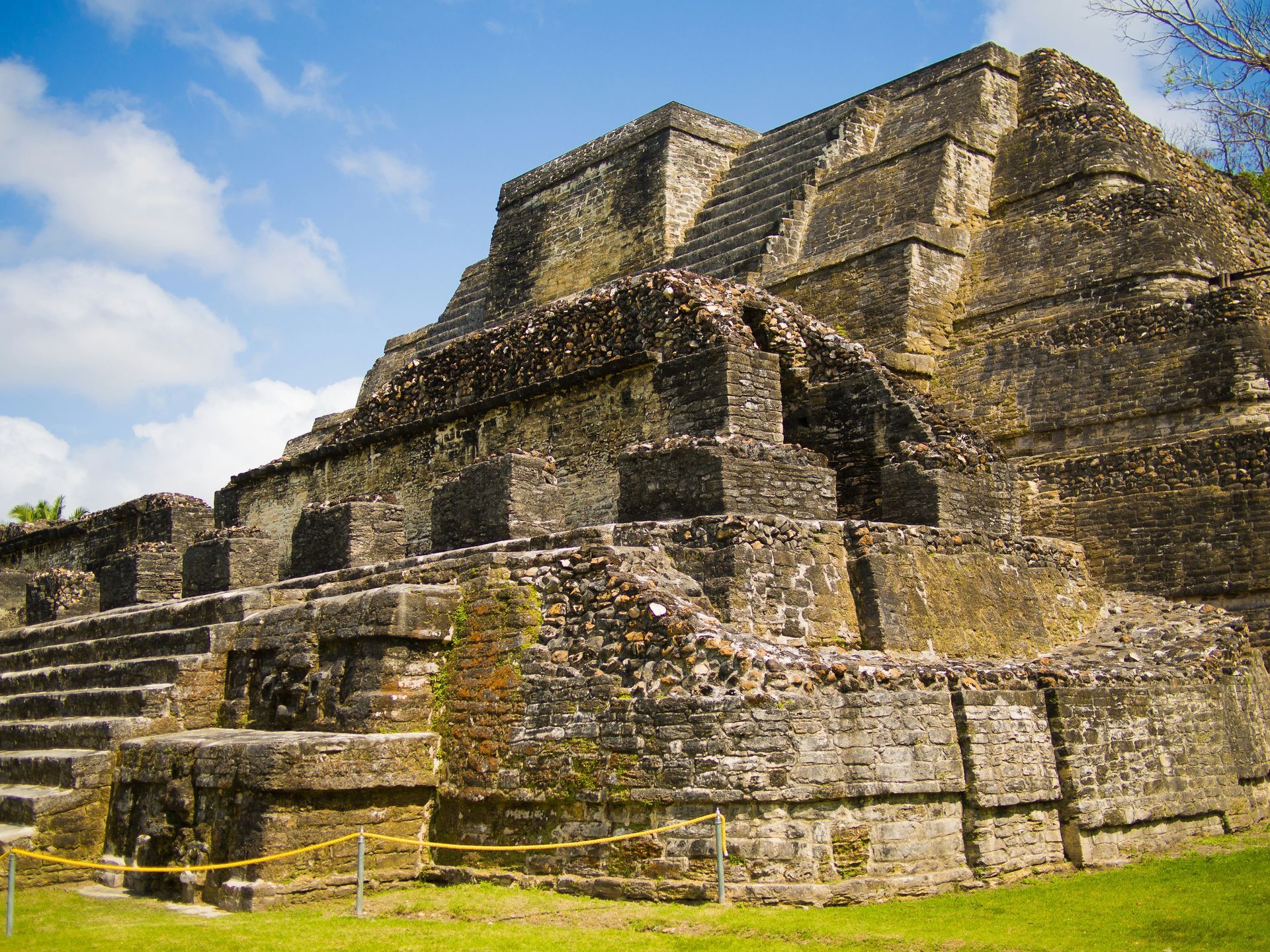 Ancient stone pyramid with stairs and weathered walls, set against a blue sky in a grassy area in Belize.