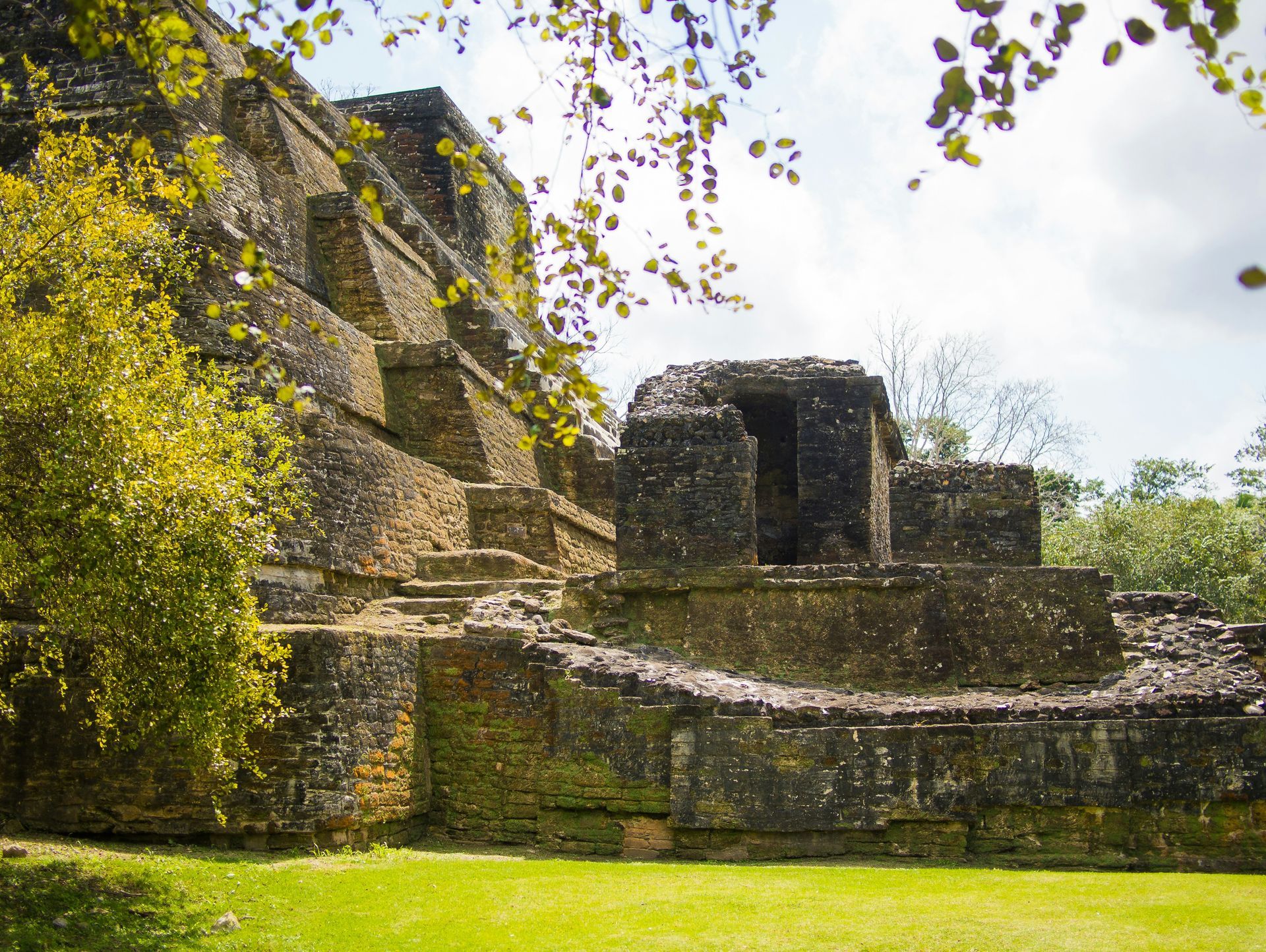 Ancient Mayan ruins, likely Xunantunich, Belize. A weathered stone structure sits amidst green grass and trees, with a cloudy sky.