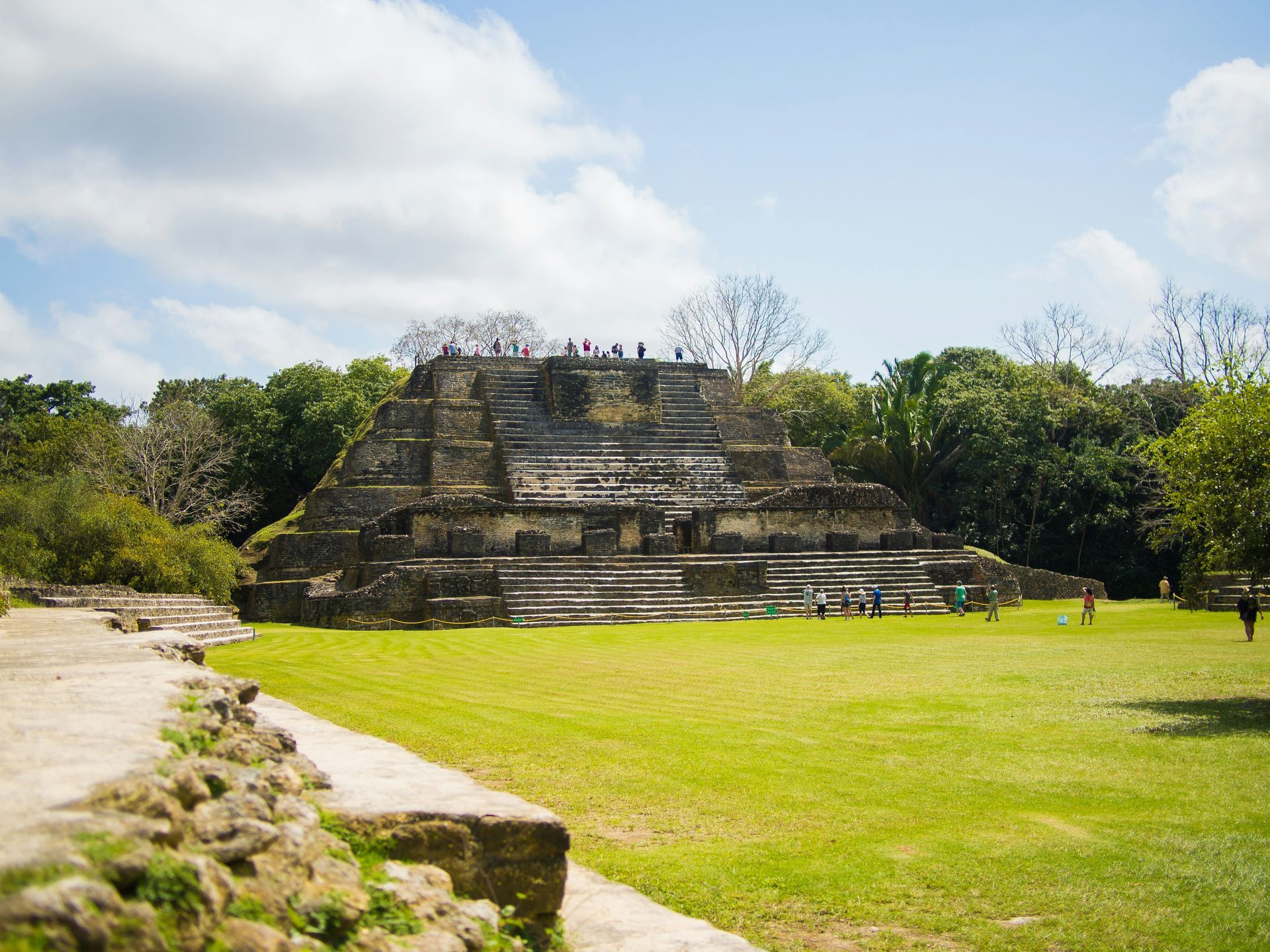Ancient Mayan temple with tiered structure on a grassy field, people exploring, surrounded by trees under a blue sky in Belize.