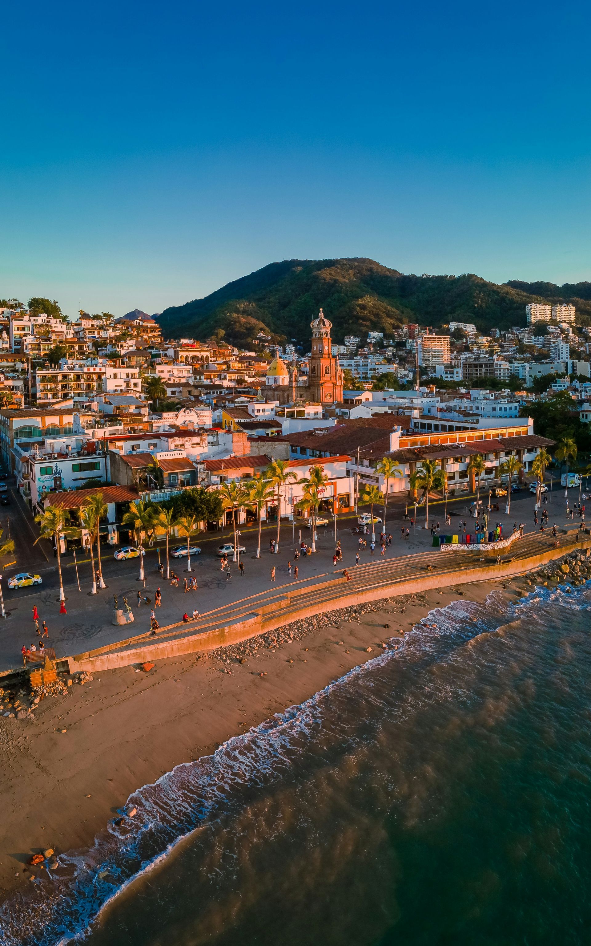 View of a Puerto Vallarta, Mexico on with buildings, a promenade, a church tower, and mountains in the background.
