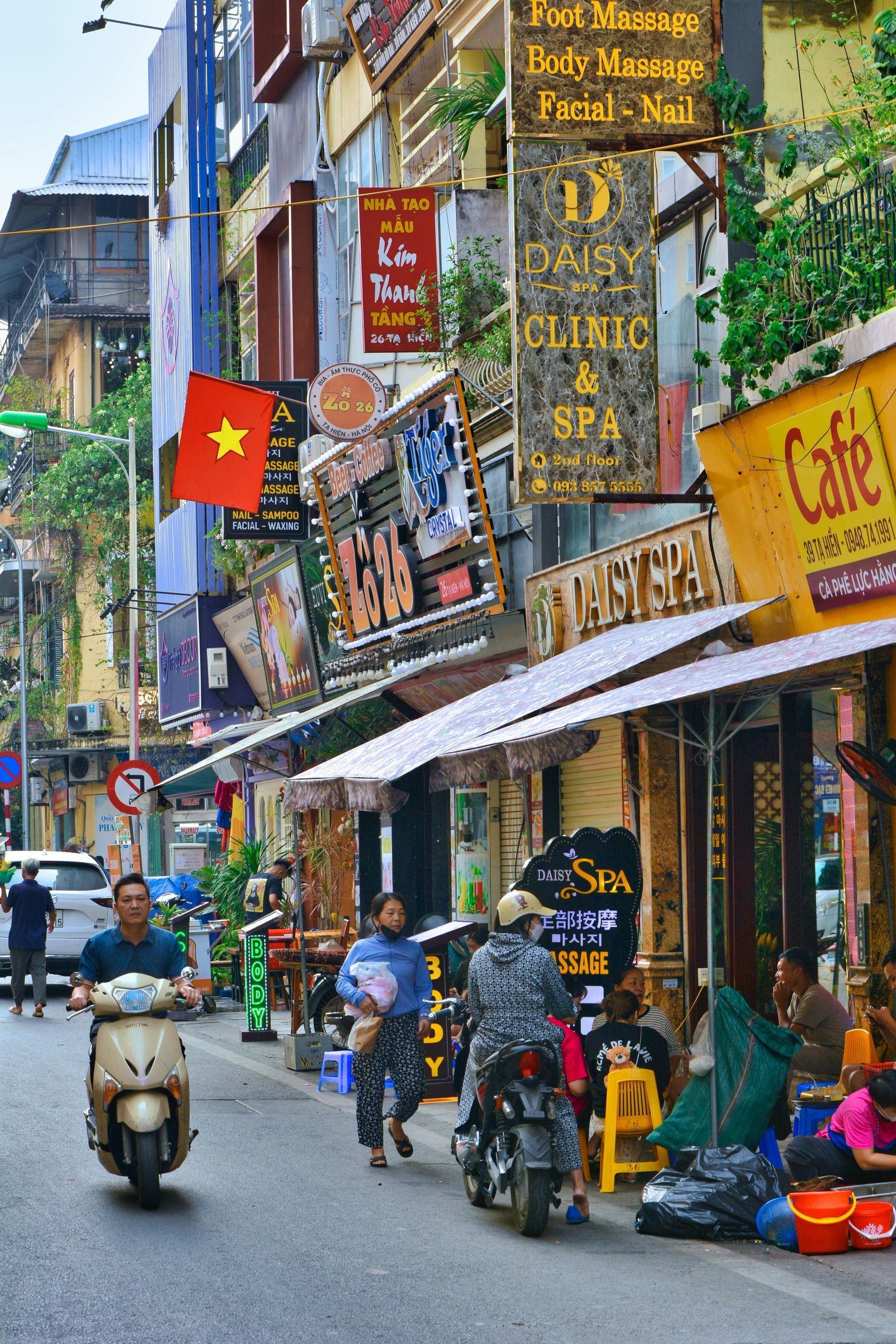 Street scene in Hanoi, Vietnam with buildings, shops, scooters, people, and Vietnamese flag.