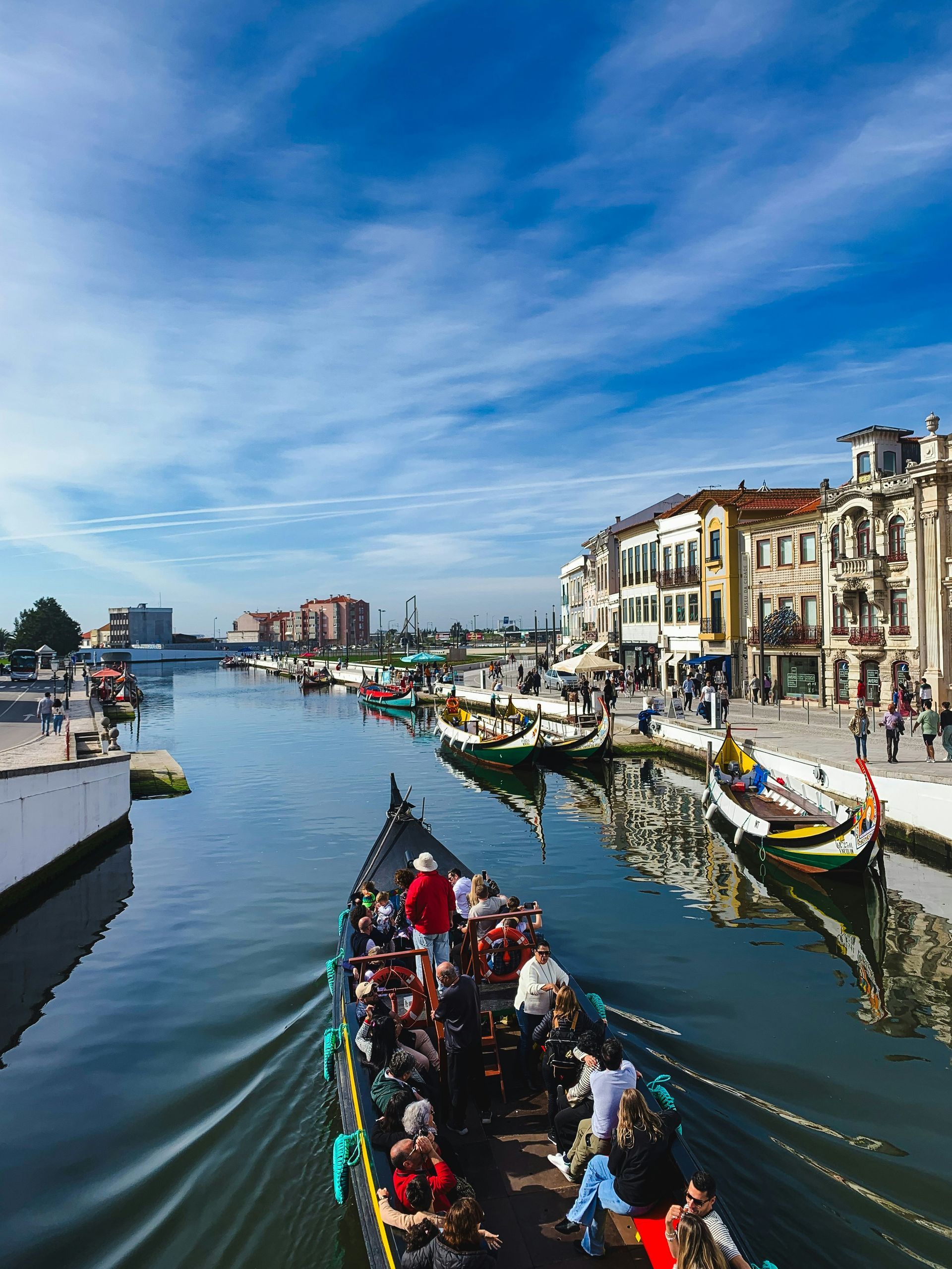 A gondola carrying tourists on a canal in Aveiro, Portugal, under a blue sky. Buildings line the canal.