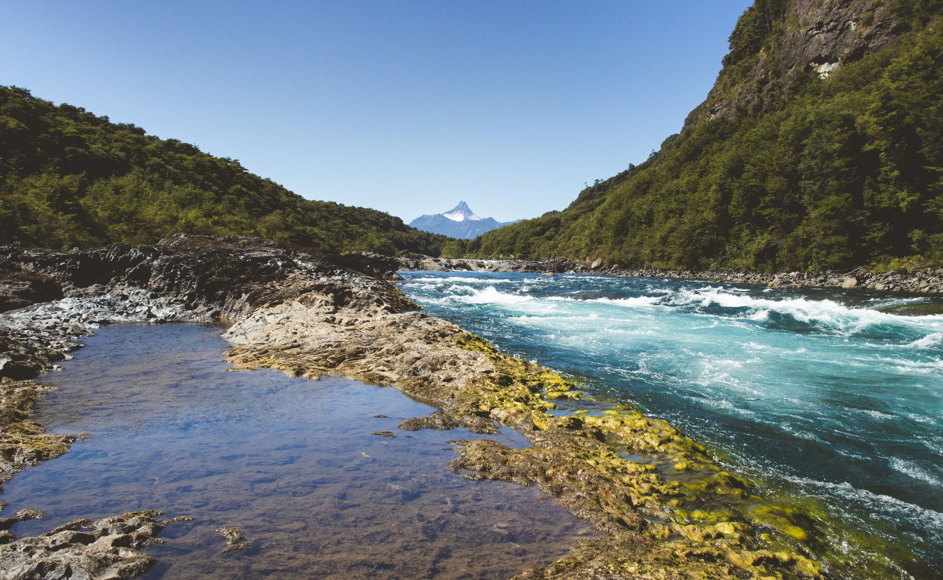 River flowing between green hills, clear blue water under a bright sky with a mountain in the distance in Los Lagos, Chile.