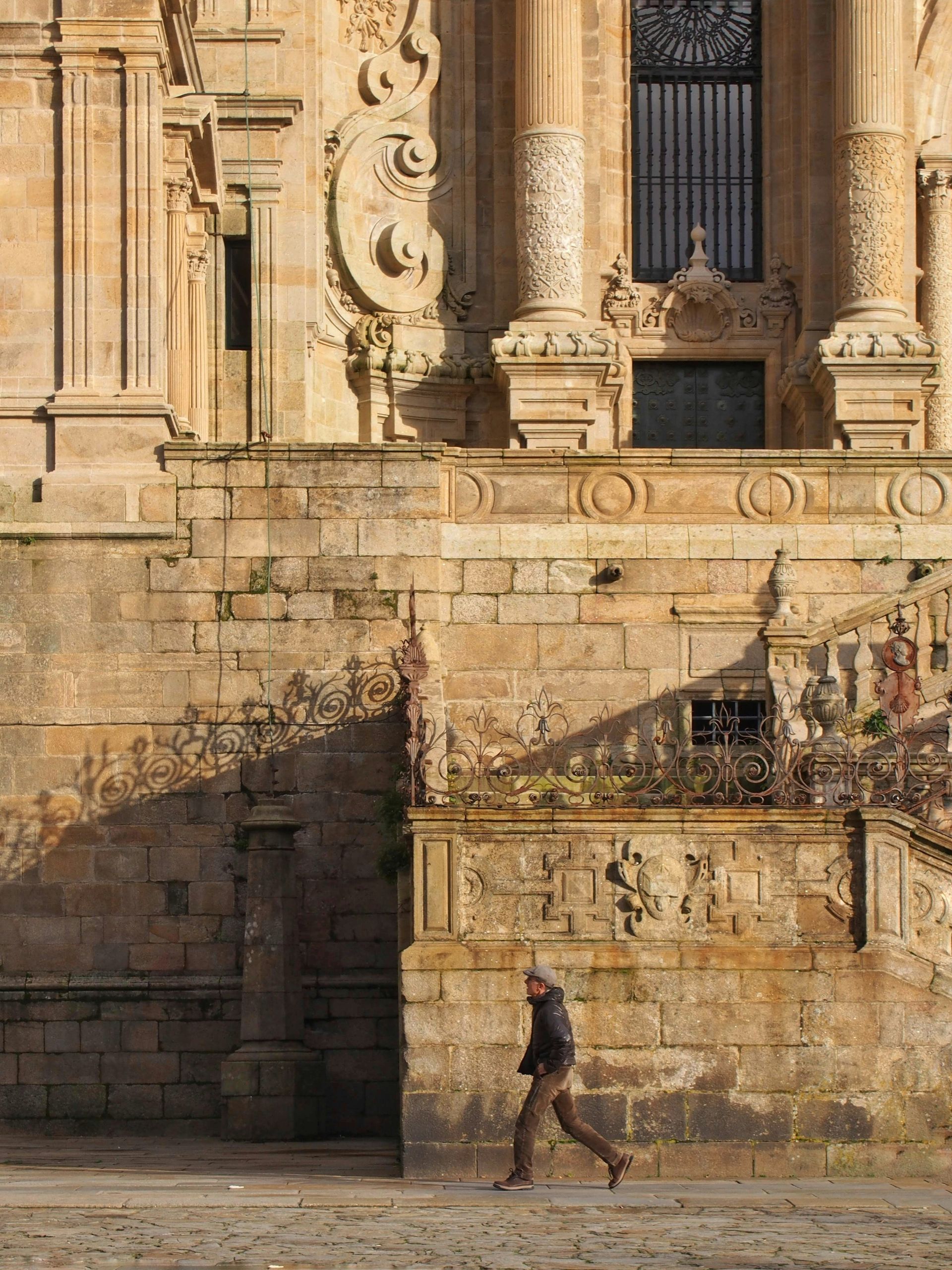Man walks past an ornate stone building with steps, likely in Santiago de Compostela, Spain.