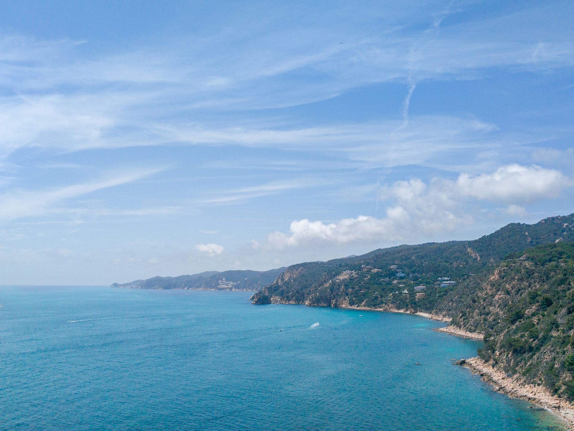 Ocean coastline with blue water, green hills, and a bright blue sky with wispy clouds on Costa Brava, Spain.