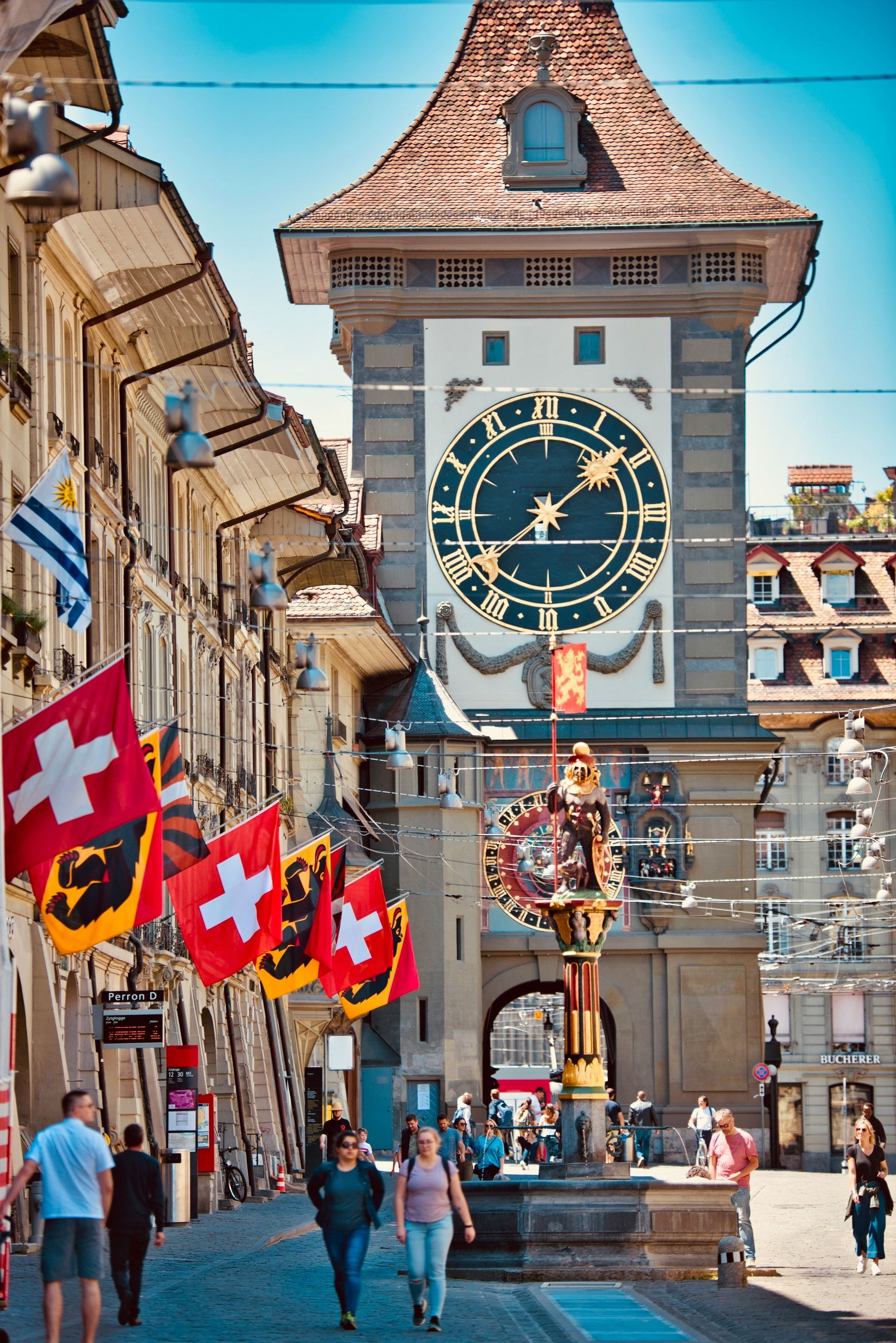 Street scene in Bern, Switzerland, with clock tower and Swiss flags. People walk on the cobblestone street.