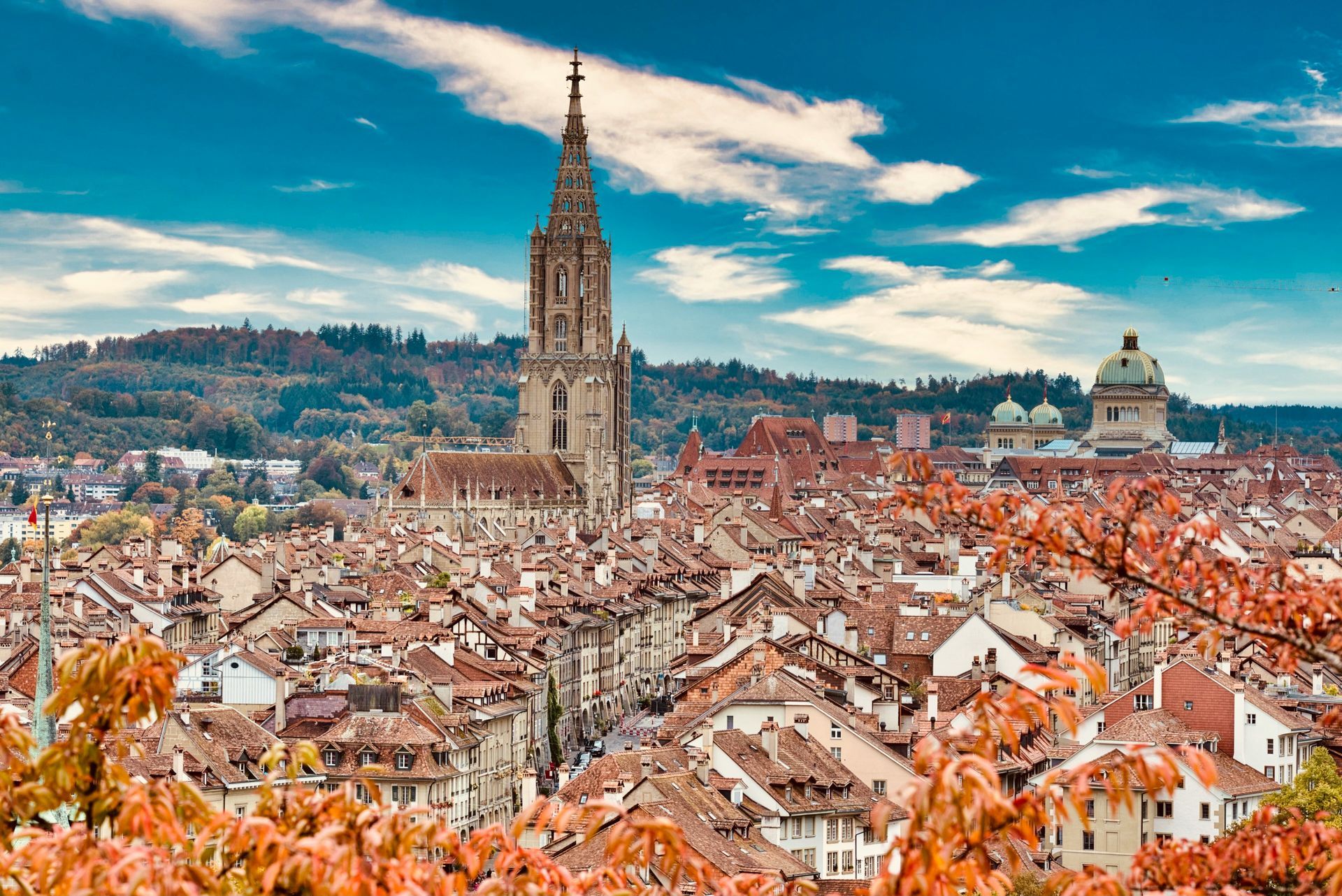 Cityscape of Bern, Switzerland, with red-tiled roofs, a tall cathedral, and vibrant fall foliage under a blue sky.