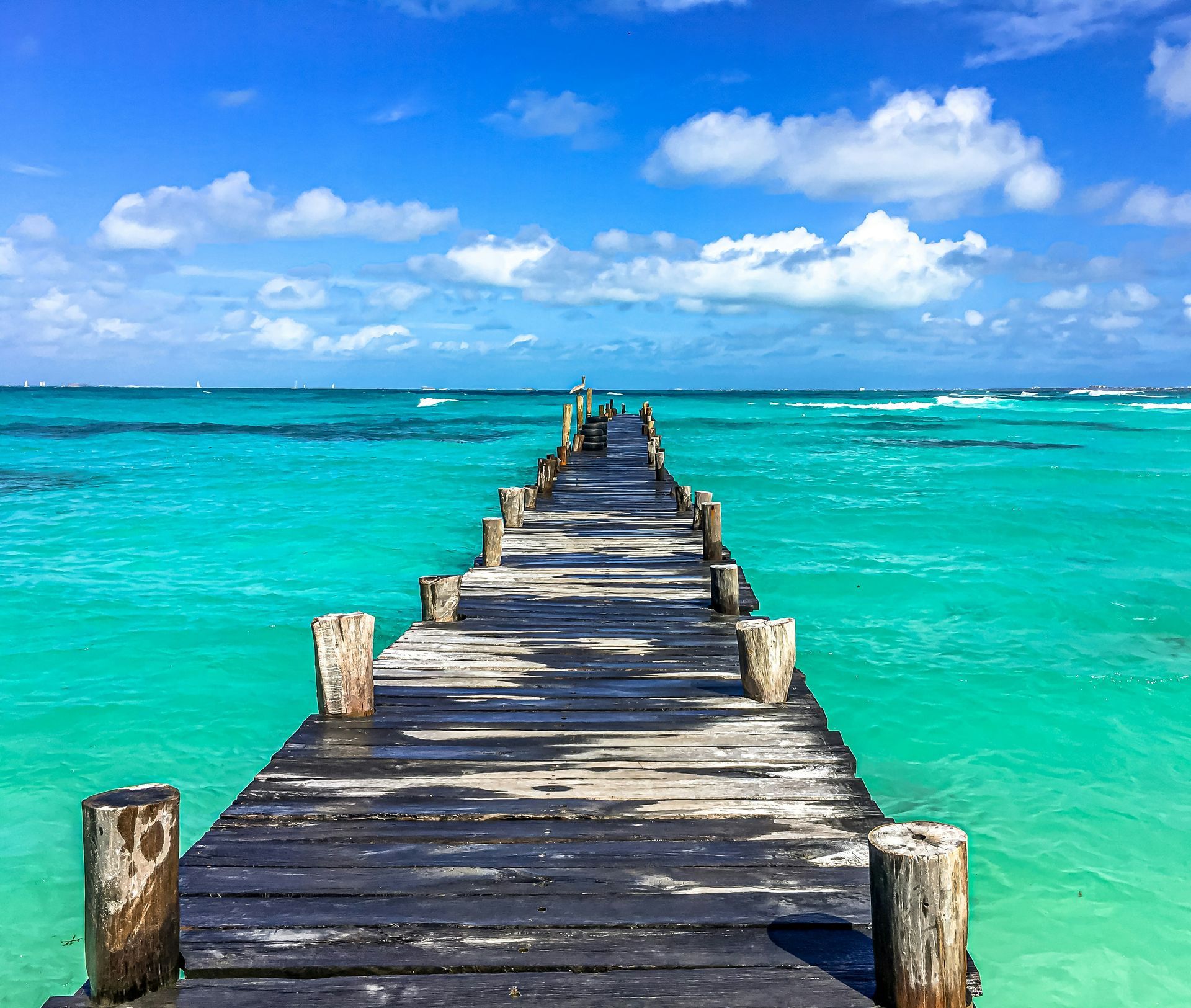 Wooden pier extends into vibrant turquoise ocean under a bright blue sky with fluffy clouds in Cancún, Mexico.