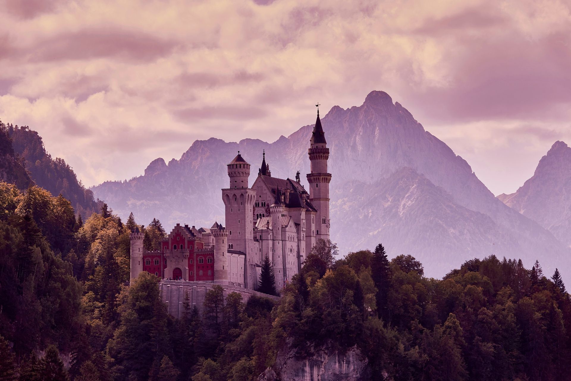 Neuschwanstein Castle atop a forested hill, silhouetted against misty mountains and a pink sky in Germany.