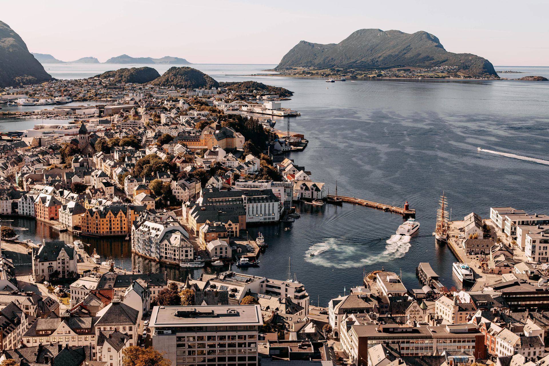 Town of Ålesund, Norway. Buildings with dark rooftops, on the water's edge. Boats on the sea and mountains in the background.