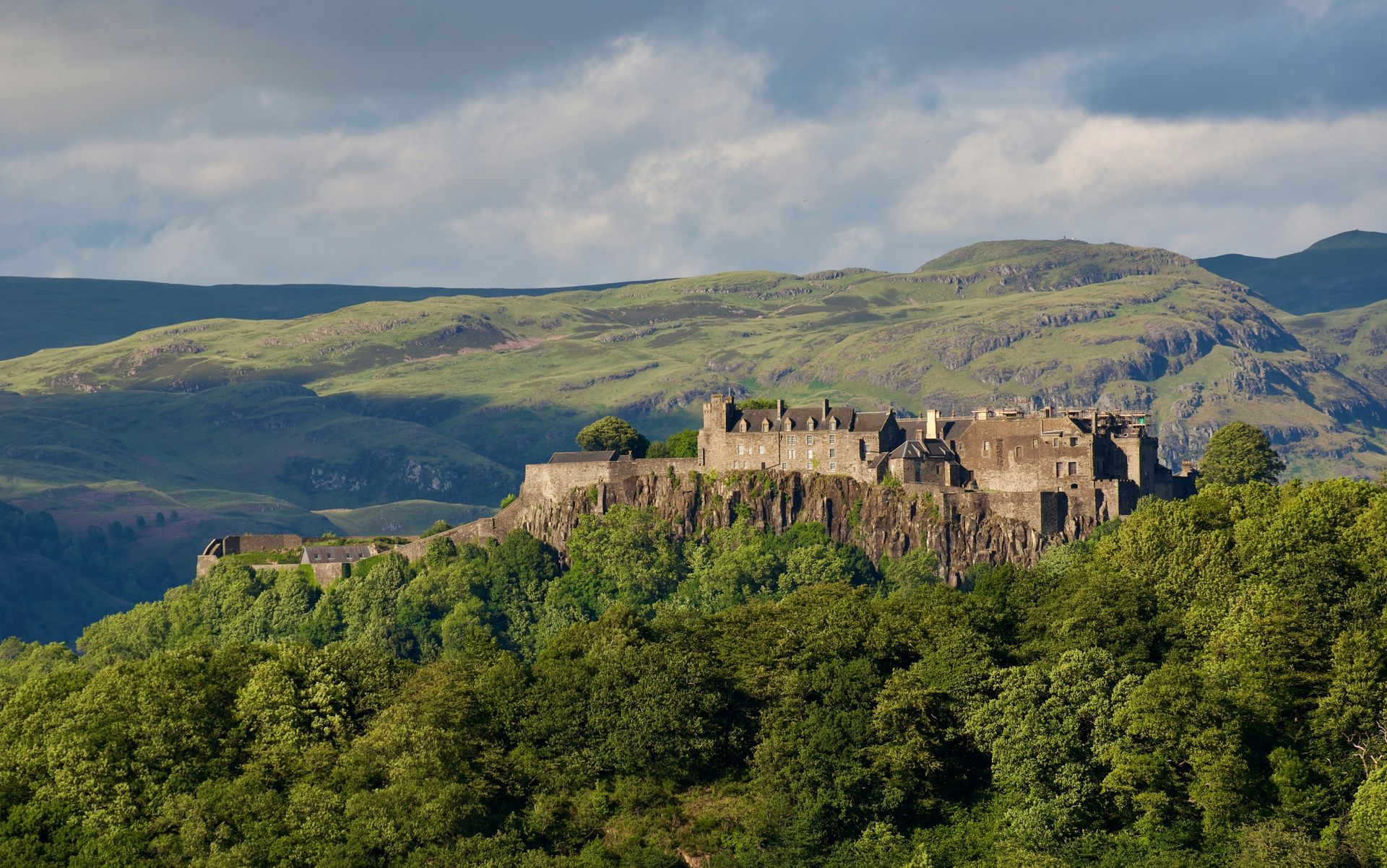 The Stirling Castle ruins atop a green, tree-covered hill with mountains in the distance under a cloudy sky in Scotland. 