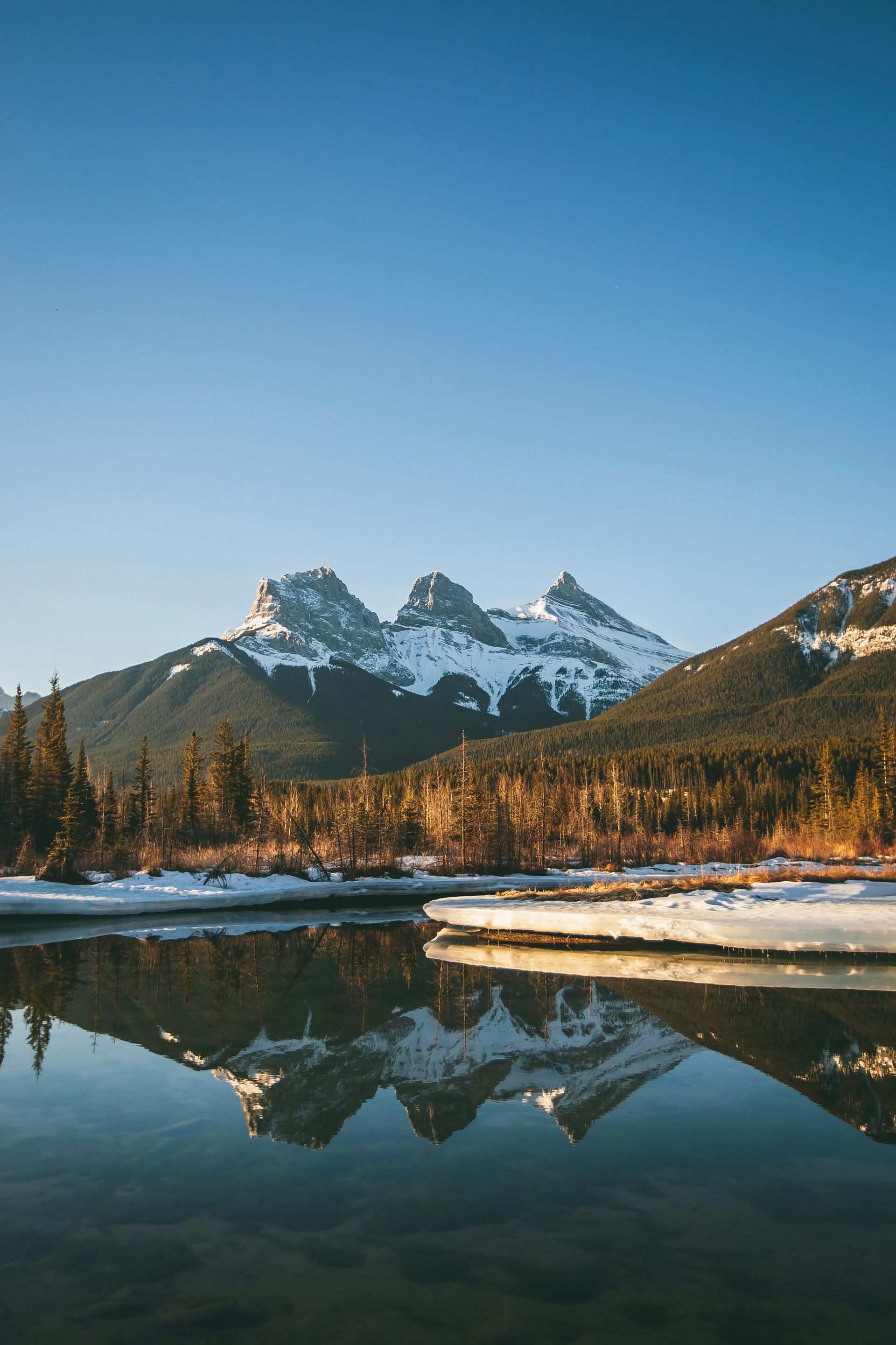 Three Sisters mountain range reflected in calm water, with snow-capped peaks and a clear blue sky in Canmore, Canada.