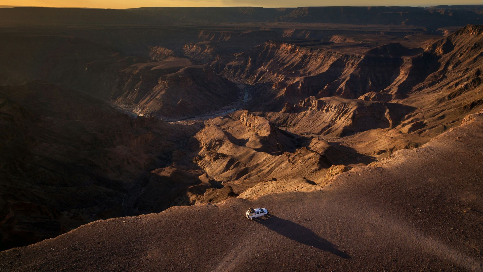 Panoramic view of Fish River Canyon in Namibia, with a vehicle on the edge under sunset.