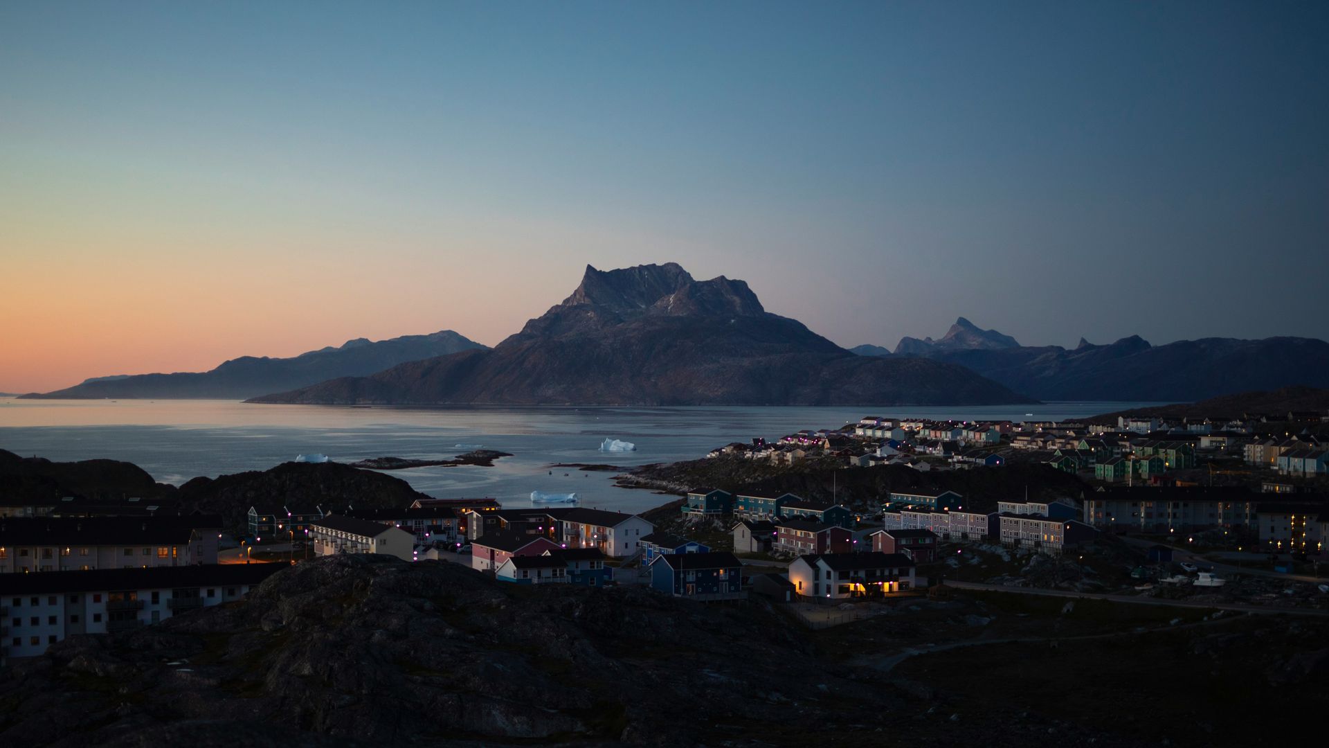 Town of Nuuk, Greenland at dusk, with illuminated buildings against a sea and mountains under a twilight sky.