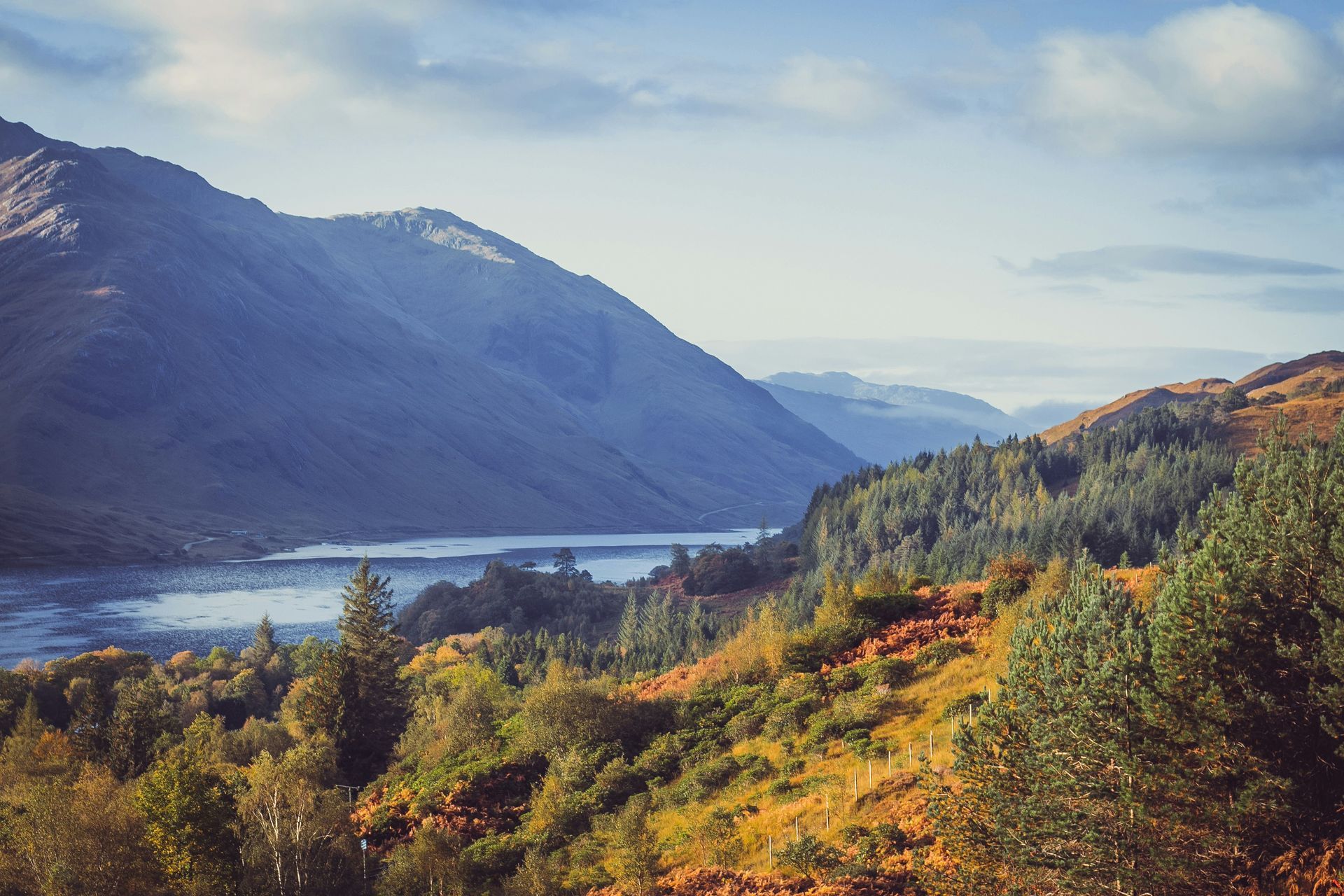 Mountains overlook Loch Shiel in Scotland, with autumn trees in the foreground and a blue sky.