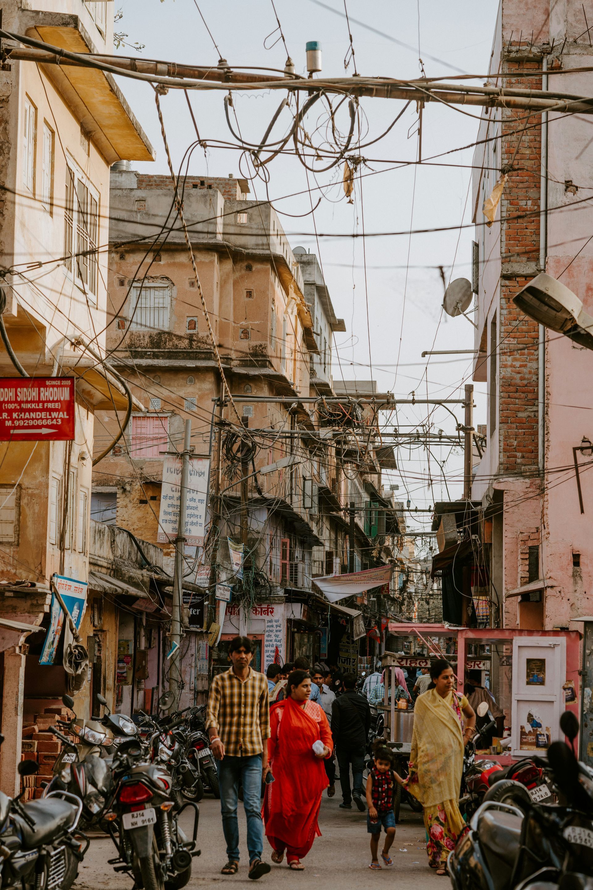 Narrow street in Jaipur, India, pedestrians walking, buildings with tangled wires overhead, motorcycles parked.