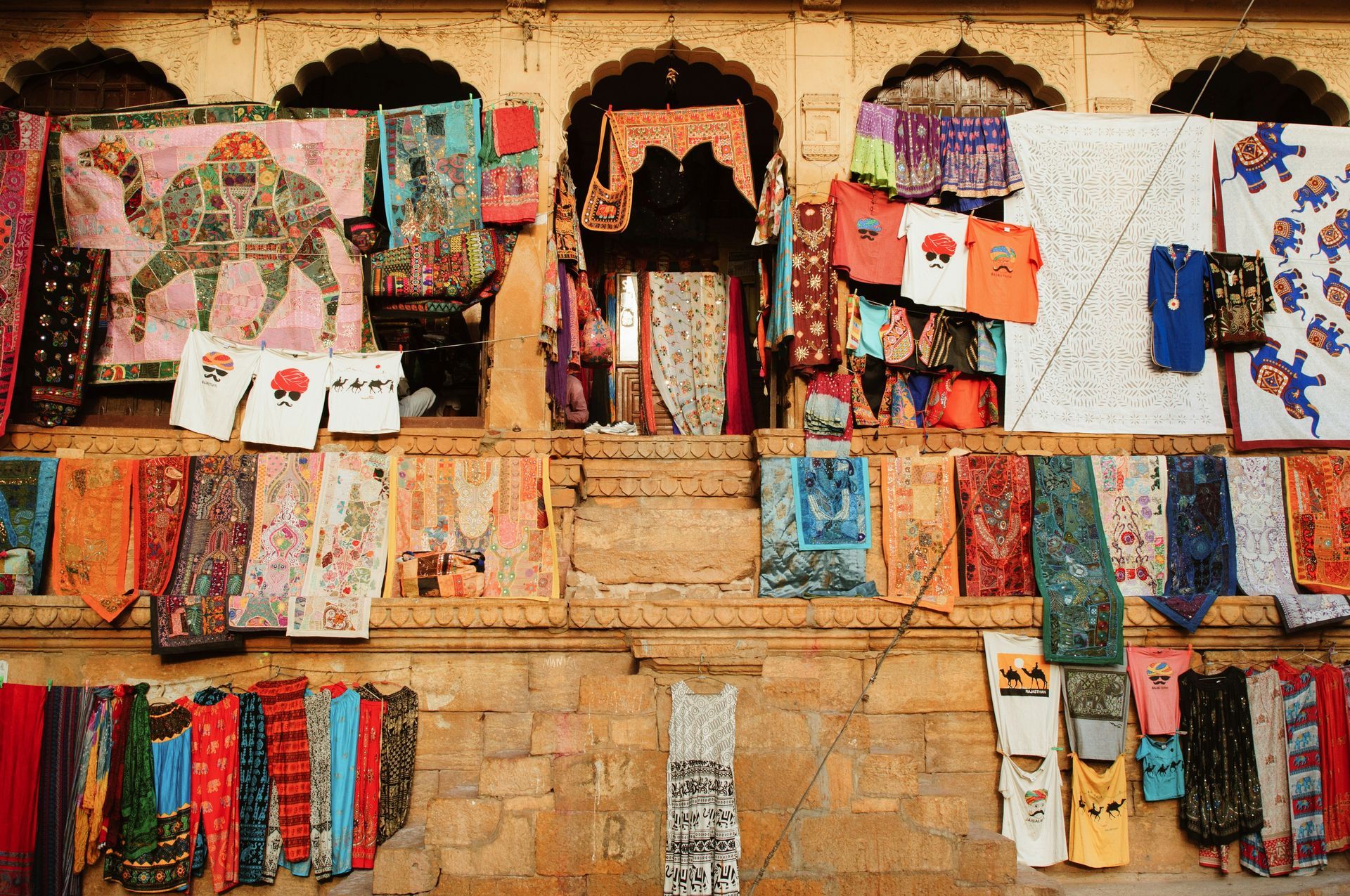 Shopfront display with colorful textiles: scarves, shirts, and cloths in a warm-toned in Jaisalmer, India.
