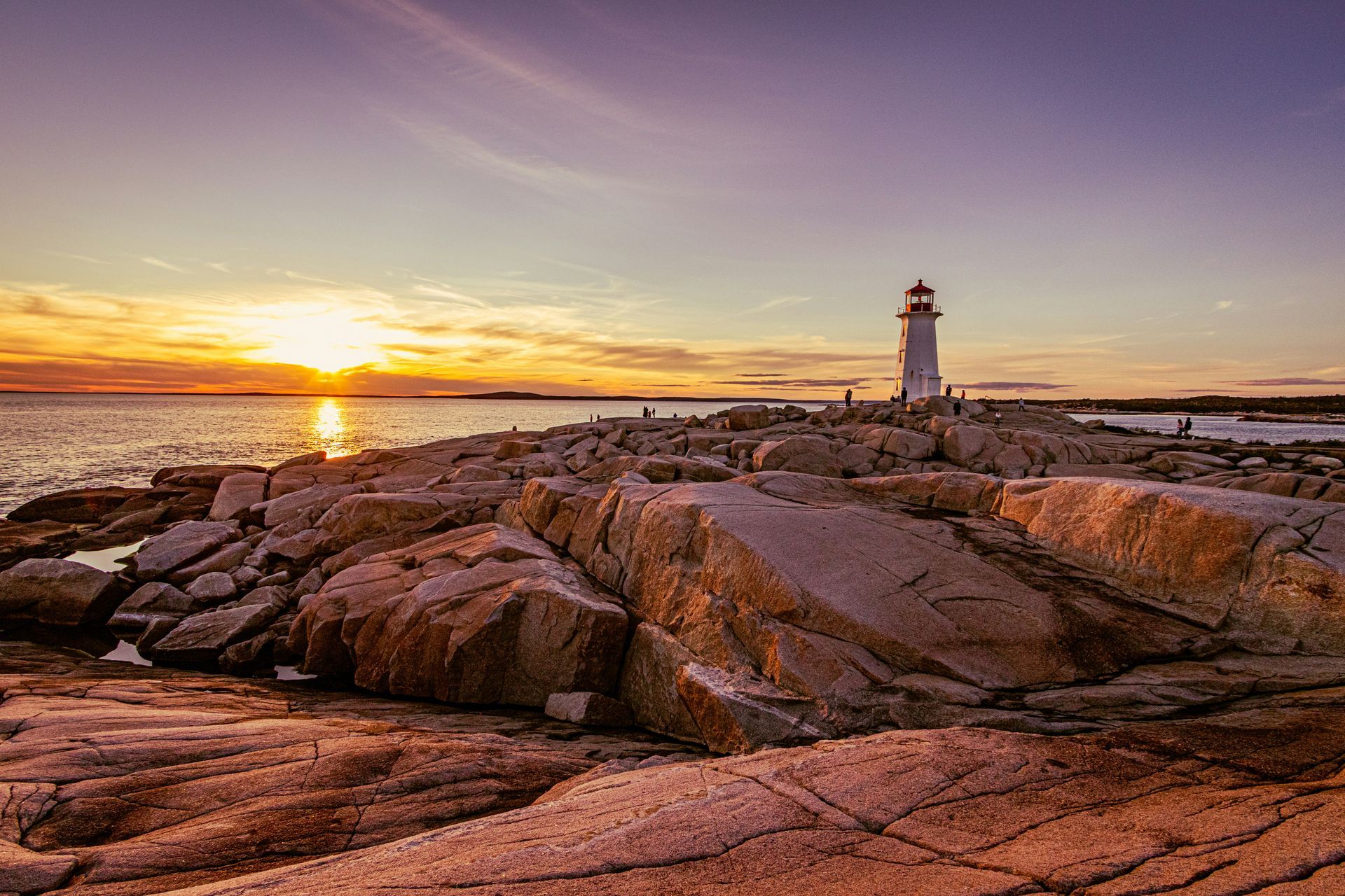 Sunset over rocky coastline with a white lighthouse. Golden light reflects off the water and rocks in Nova Scotia, Canada.