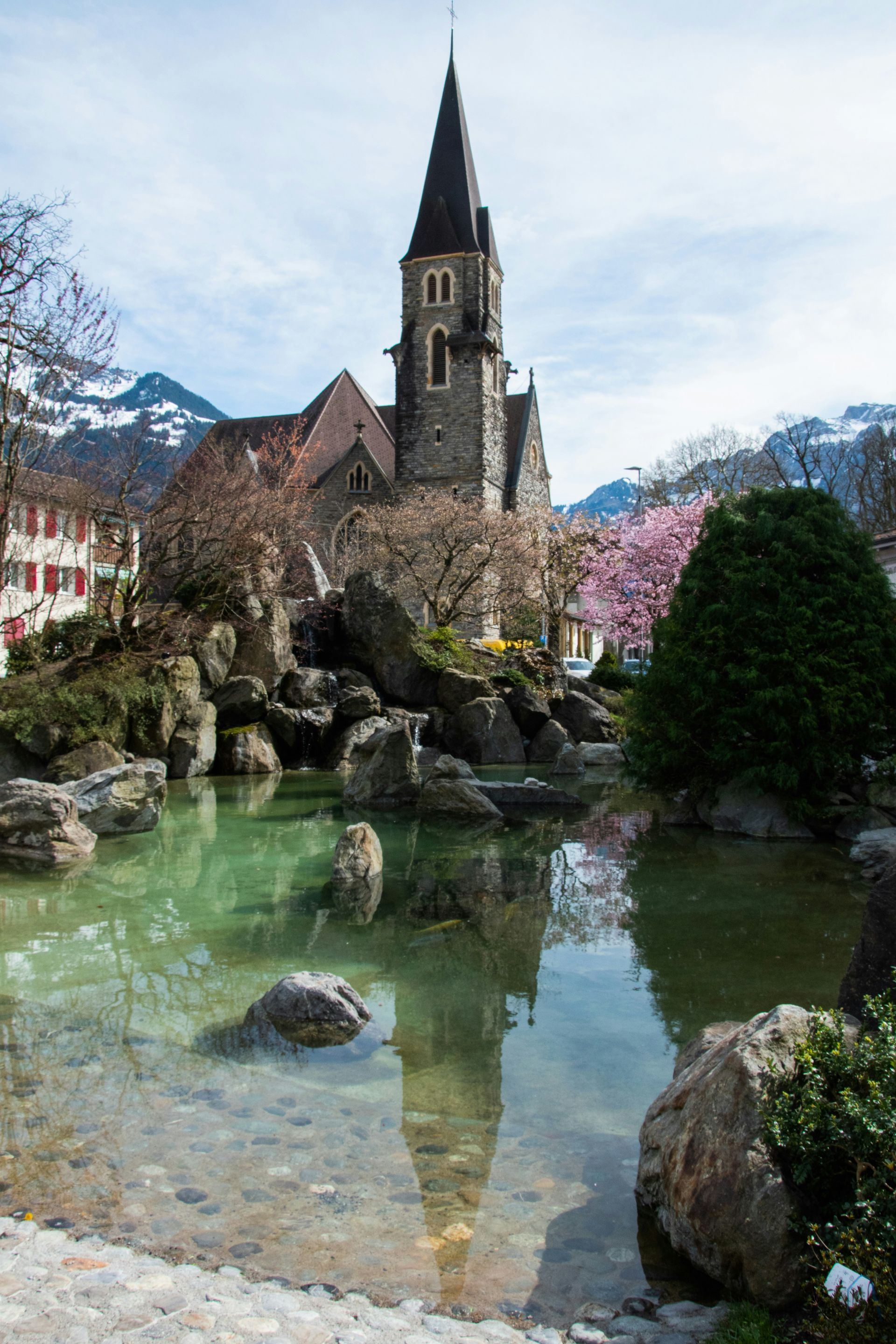 Church with a tall steeple reflected in a pond with rocks, surrounded by trees and mountains in Interlaken, Switzerland.