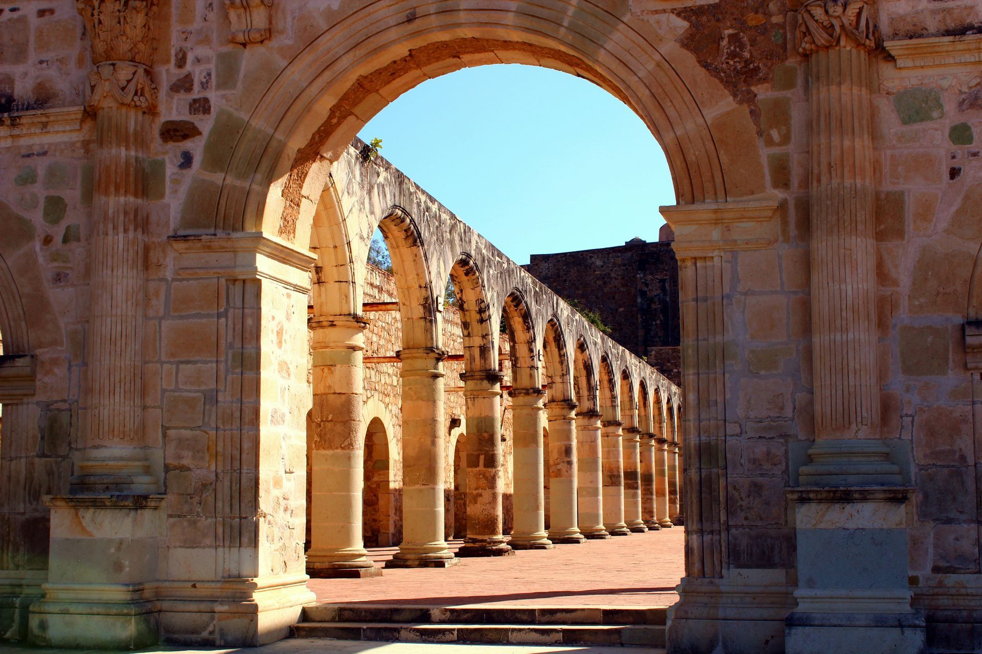 Stone archway framing a row of arched columns in a sunlit, weathered building; blue sky visible in Oaxaca, Mexico.