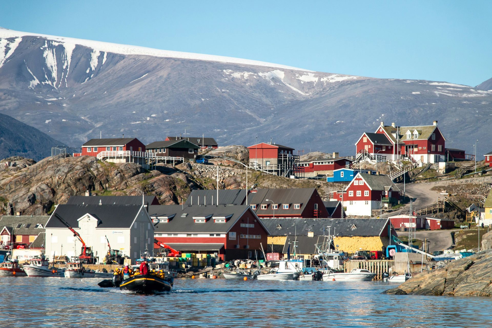 Colorful buildings on rocky shoreline; boats in water, snow-capped mountains in the background in Uummannaq, Greenland.