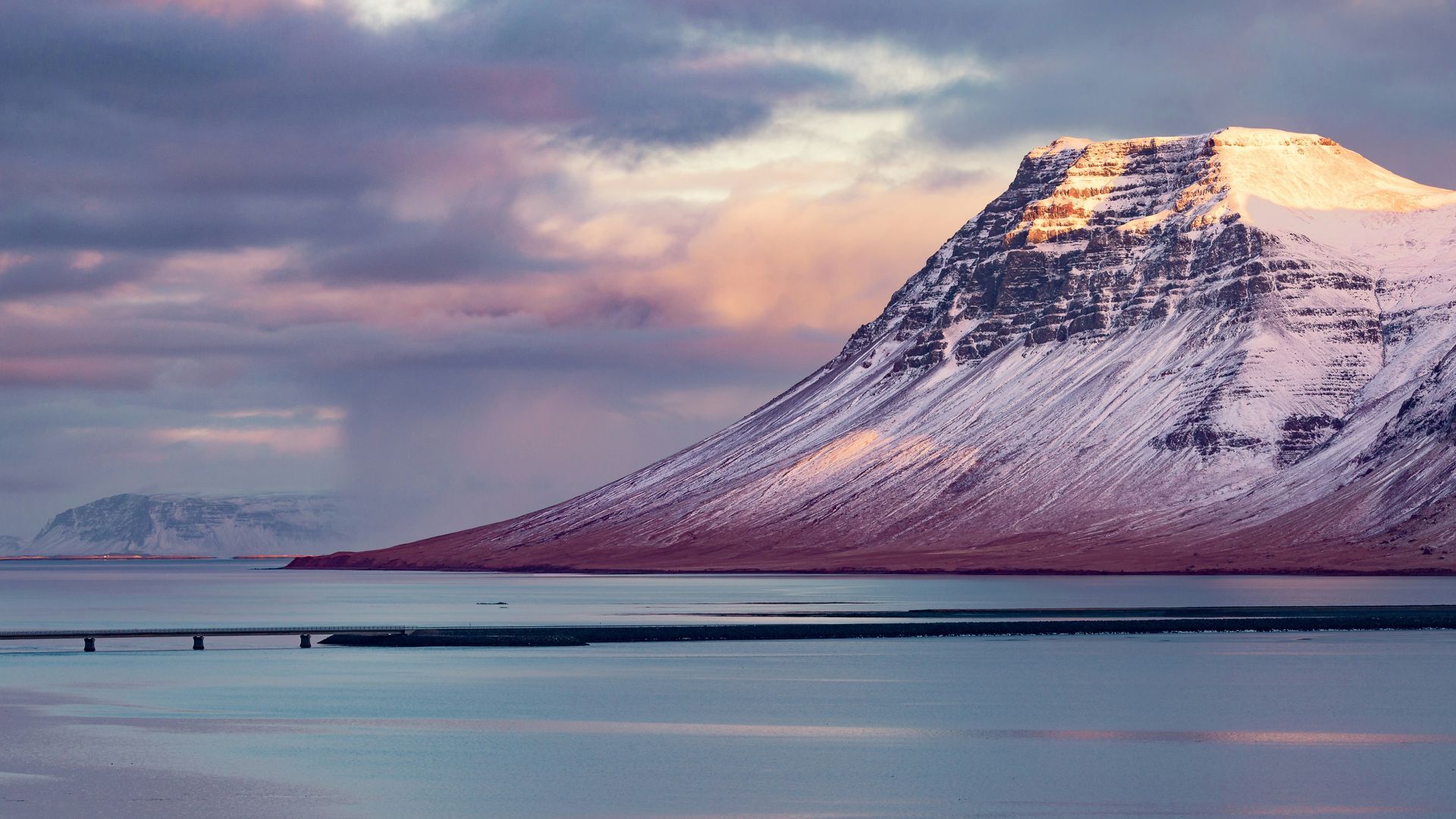 Snow-capped mountain with golden sunlight, reflected in calm water with a bridge in the foreground, purple and pink sky in Snæfellsnes, Iceland.