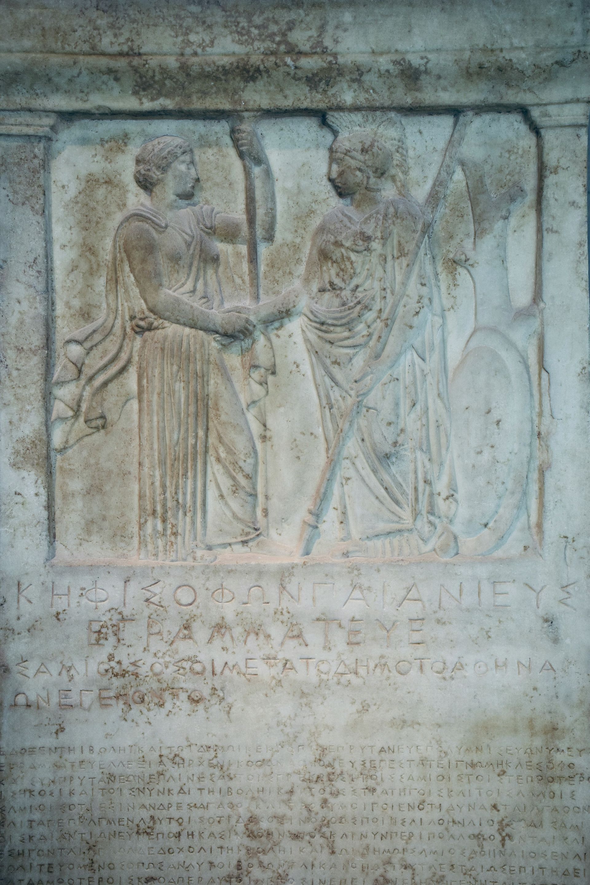 A statue of two women standing next to each other on a stone wall in Greece.