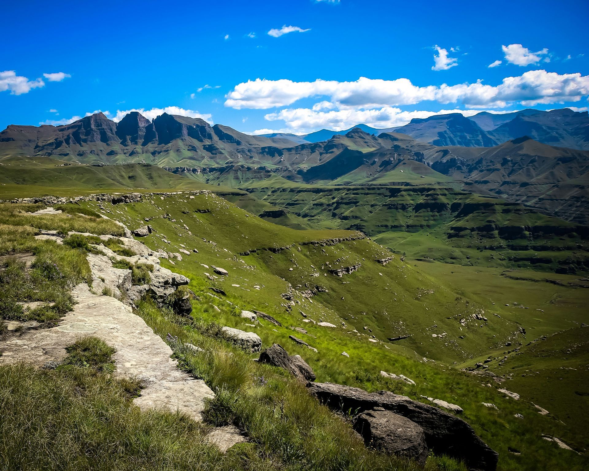 Green hills and valleys stretch toward a rugged mountain range in the Drakensberg, South Africa.