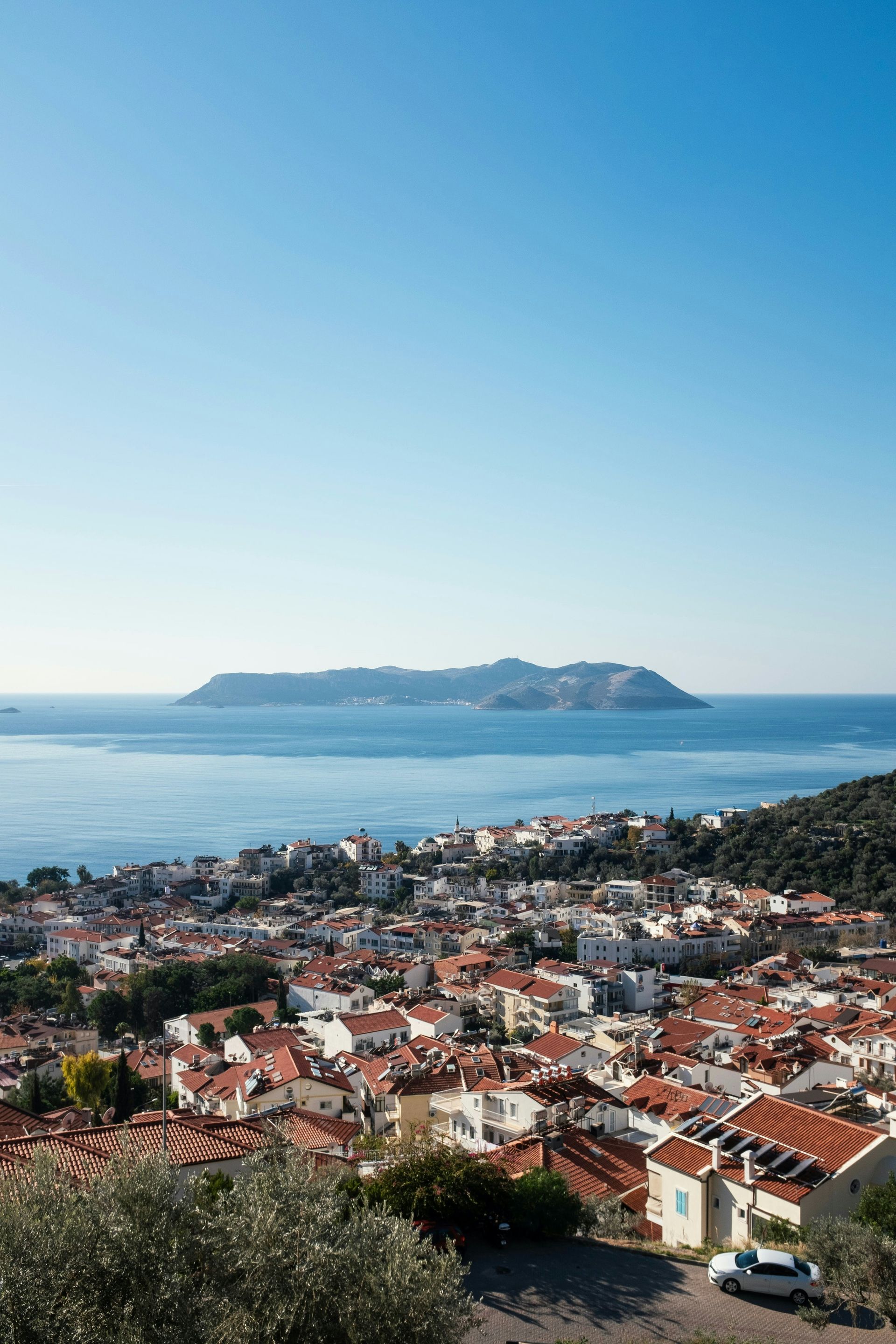 Kaş, Turkey, overlooking a blue sea under a clear sky. Red-roofed buildings, a distant island.