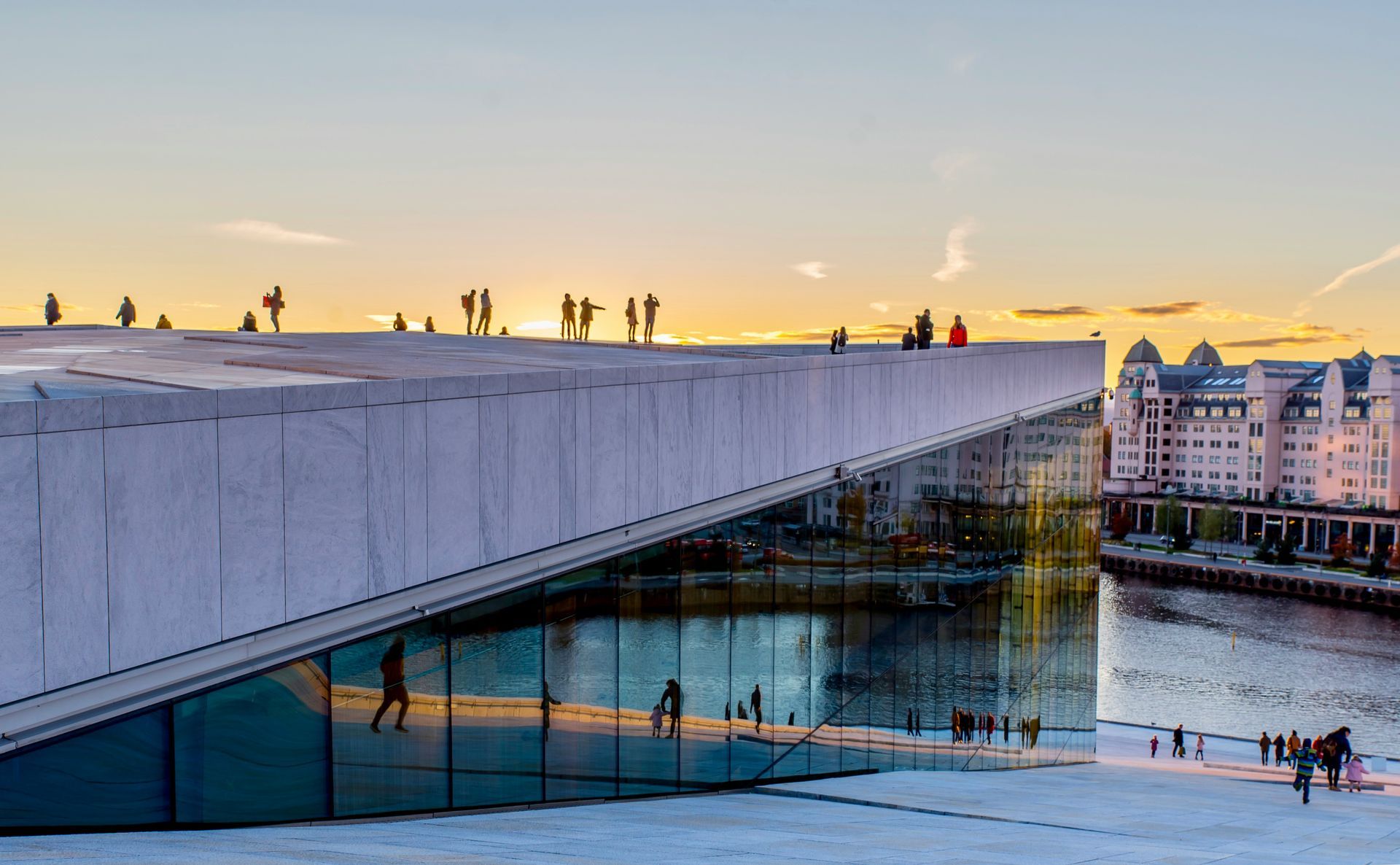 People on roof of Oslo Opera House, sunset reflections on glass and water, modern architecture.