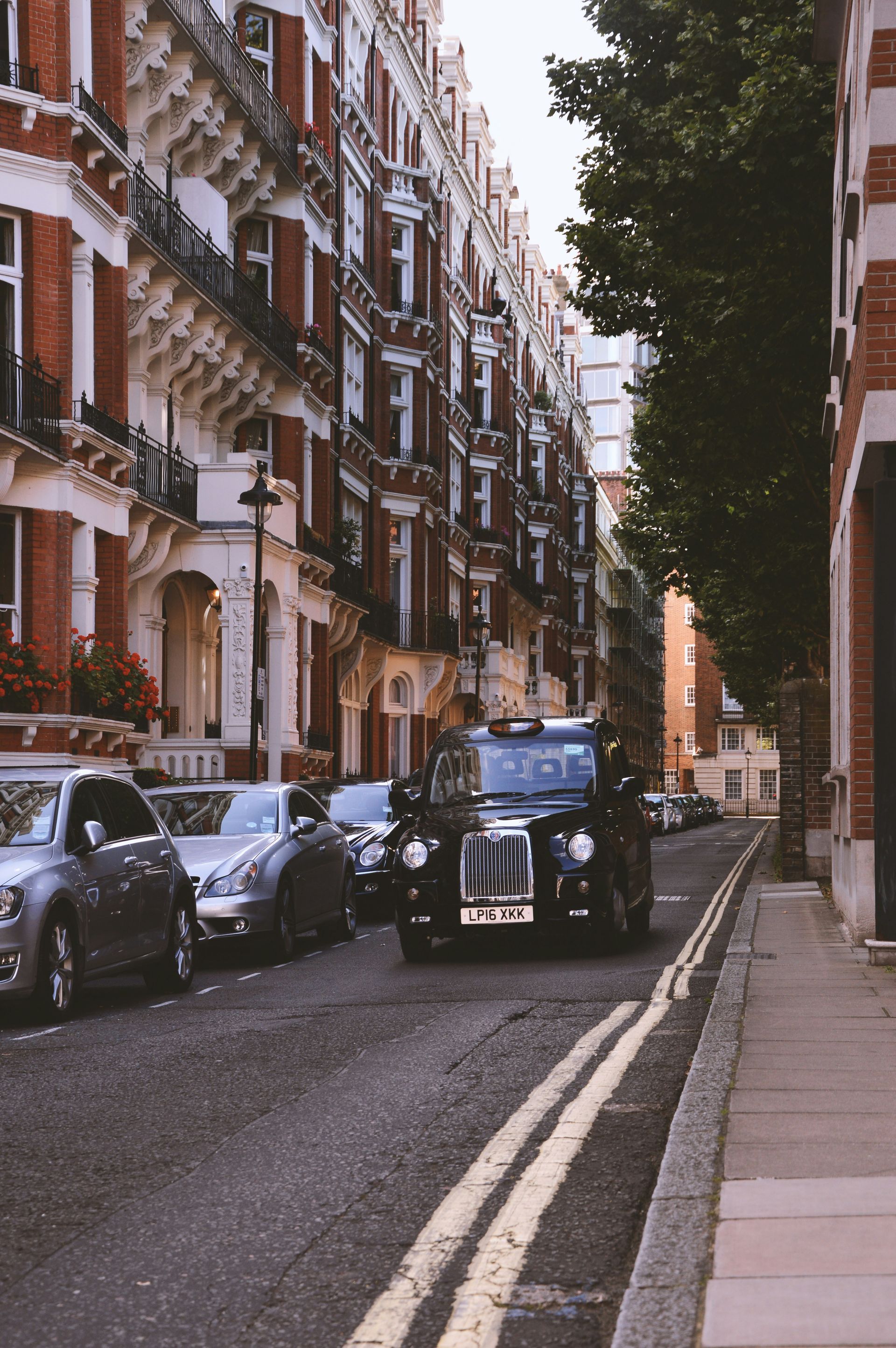 London street scene: Black taxi driving, parked cars, ornate buildings with red brick, a tree, and sidewalk.