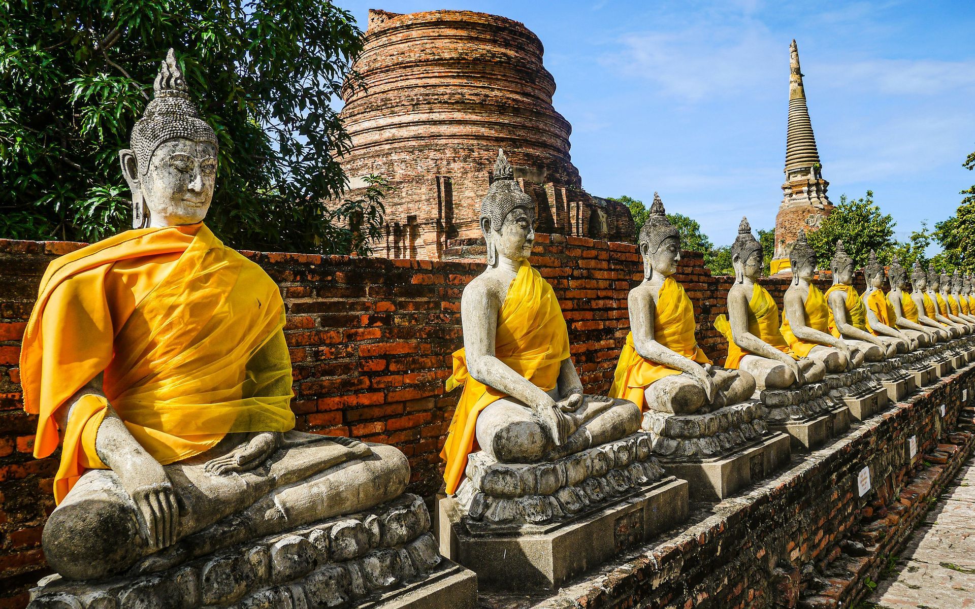 A line of Buddha statues with yellow robes are sitting in front of the ruins of Ayutthaya, Thailand.