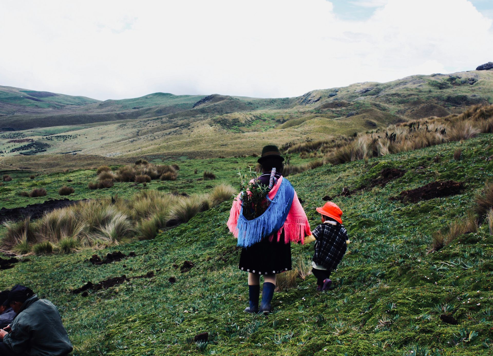Woman in traditional garb and child walk up a grassy hillside, mountains in the distance in Ecuador.