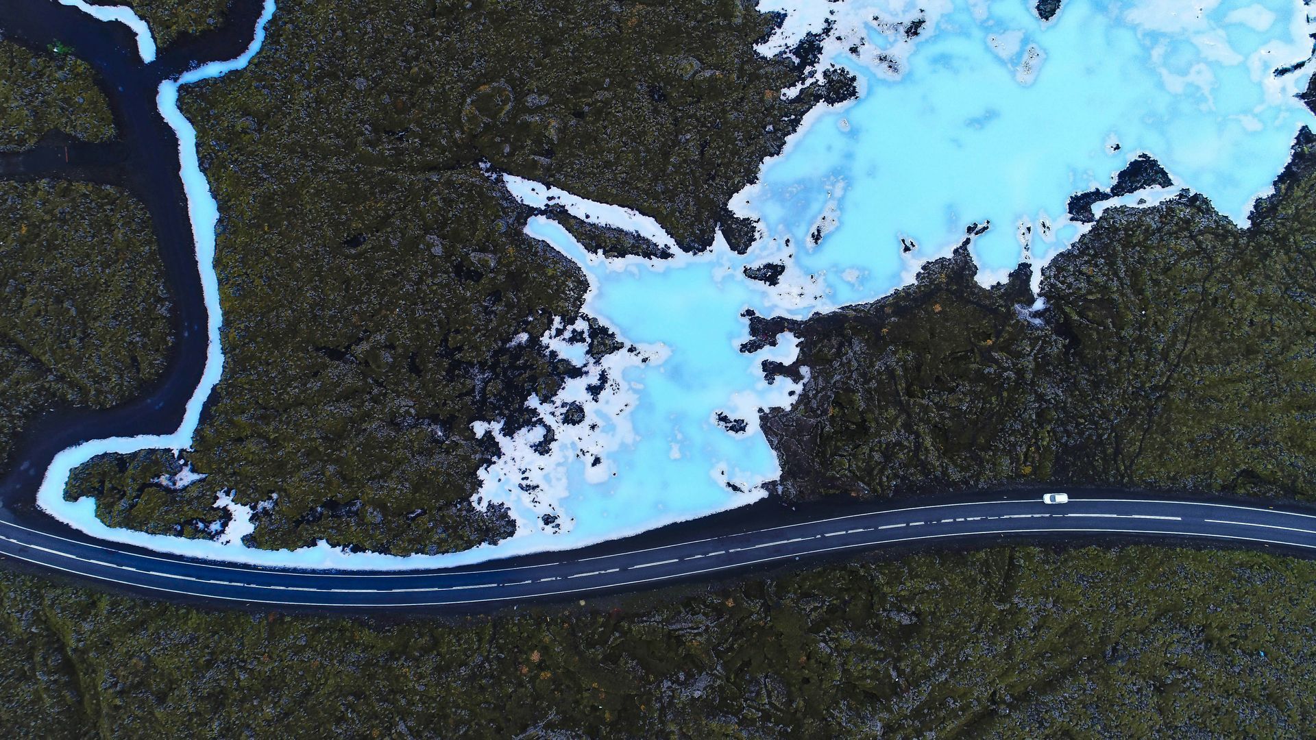 Road winds through dark foliage and turquoise waters at Blue Lagoon, Iceland.