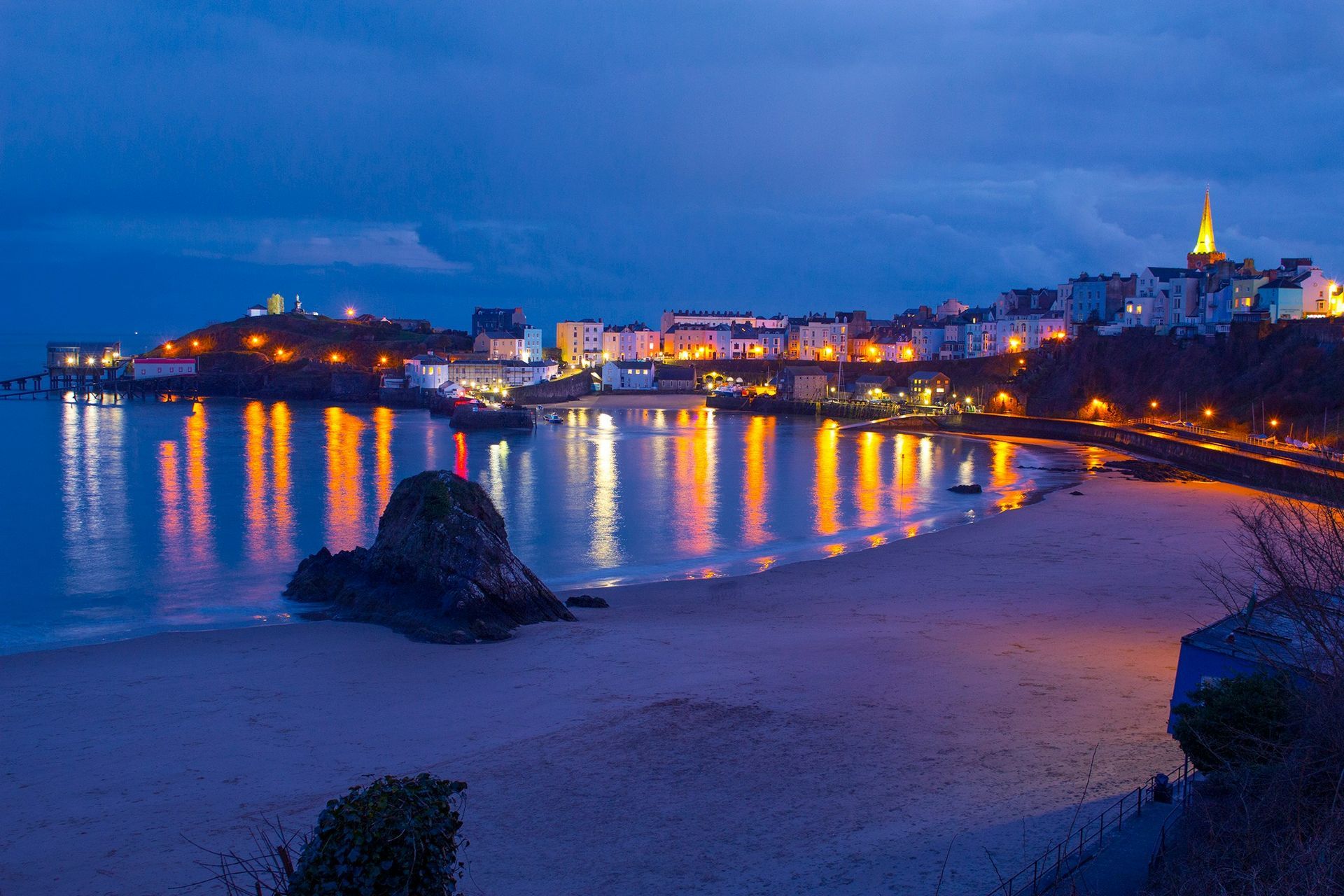 Night view of Tenby, Wales, with illuminated buildings reflected in the calm water of the beach.