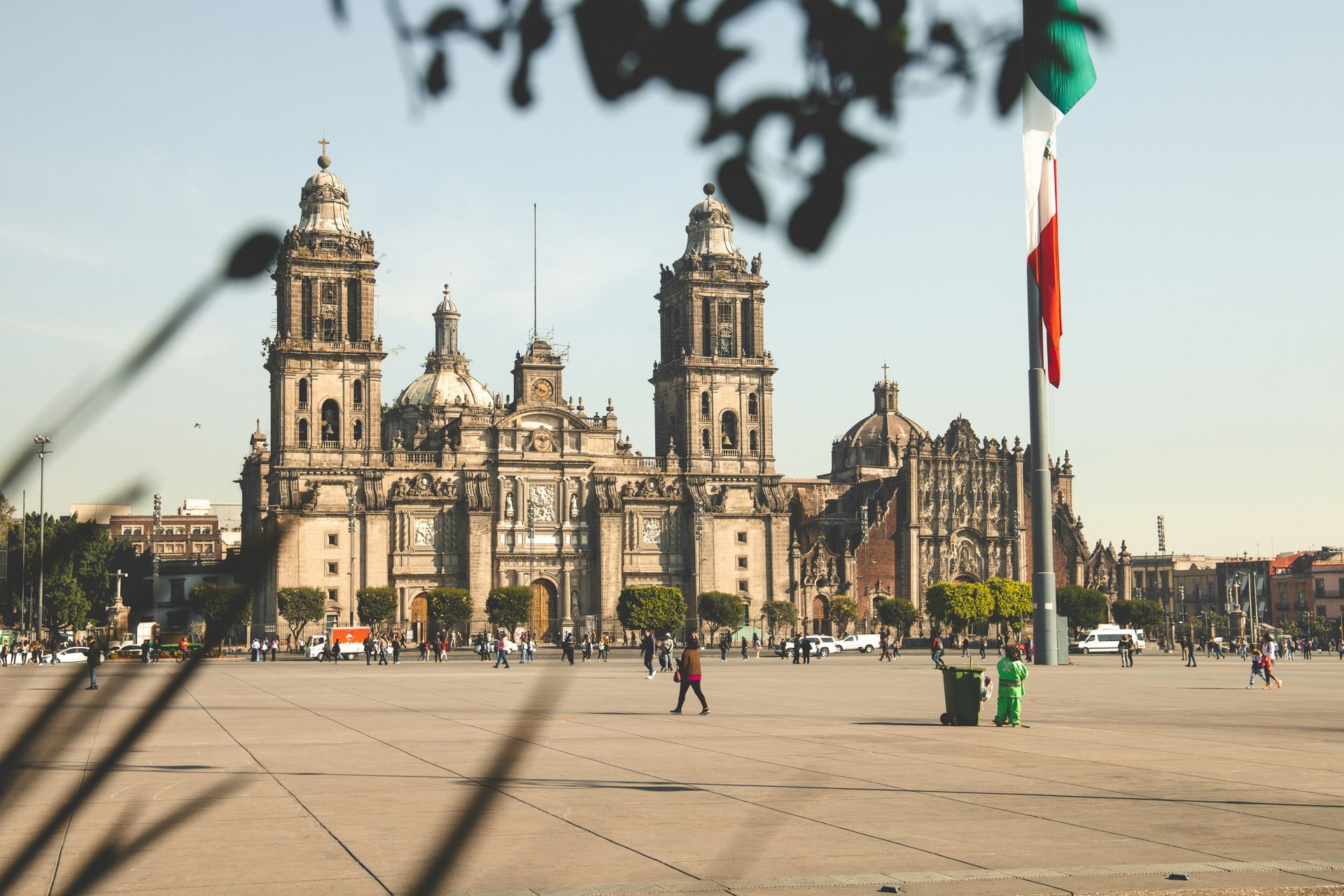 Mexico City's Metropolitan Cathedral on a sunny day, with people walking in the square and the Mexican flag flying.