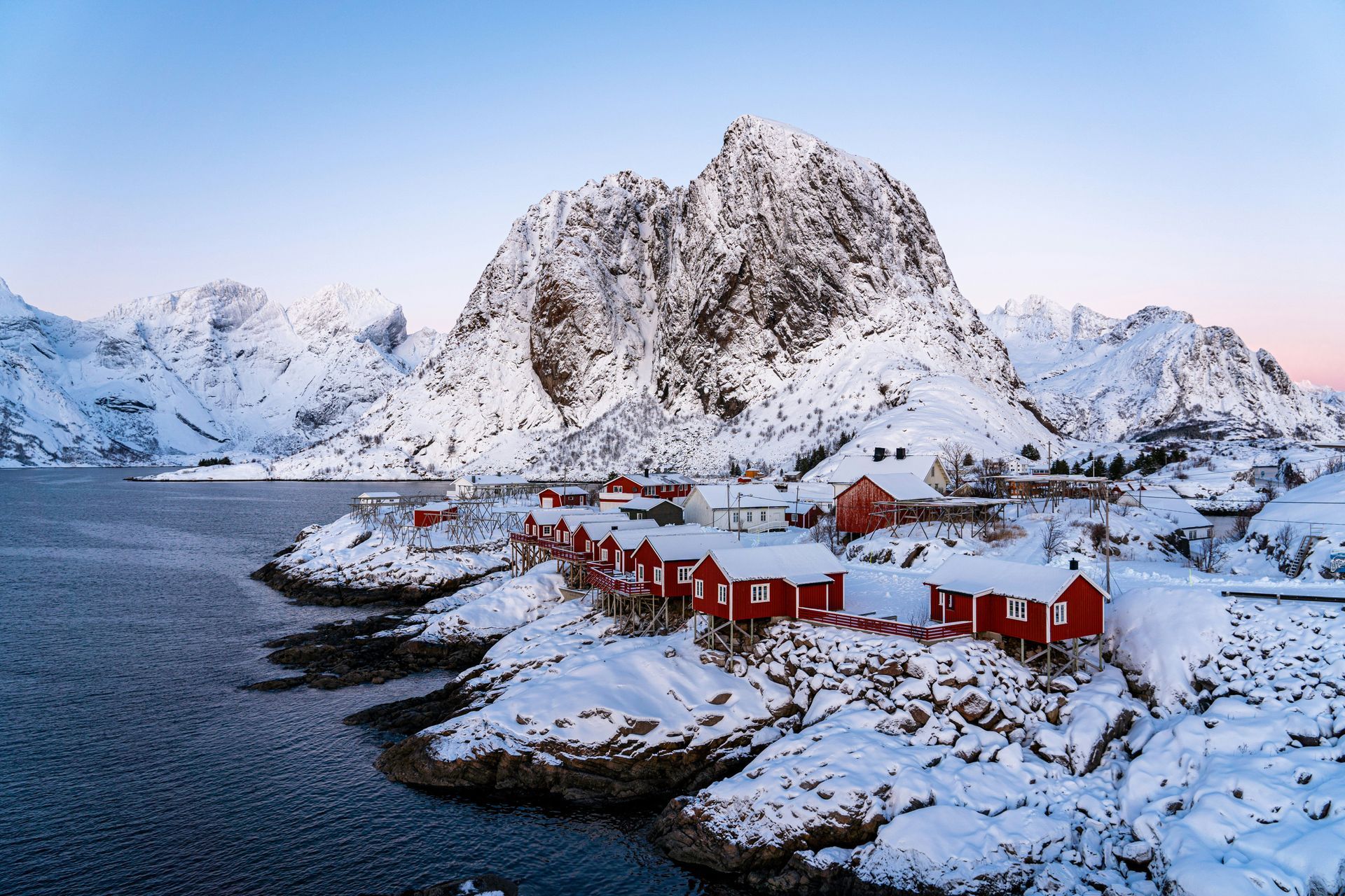 Red cabins line a snow-covered shore at the base of a snow-capped mountain, on the ocean in Lofoten, Norway.