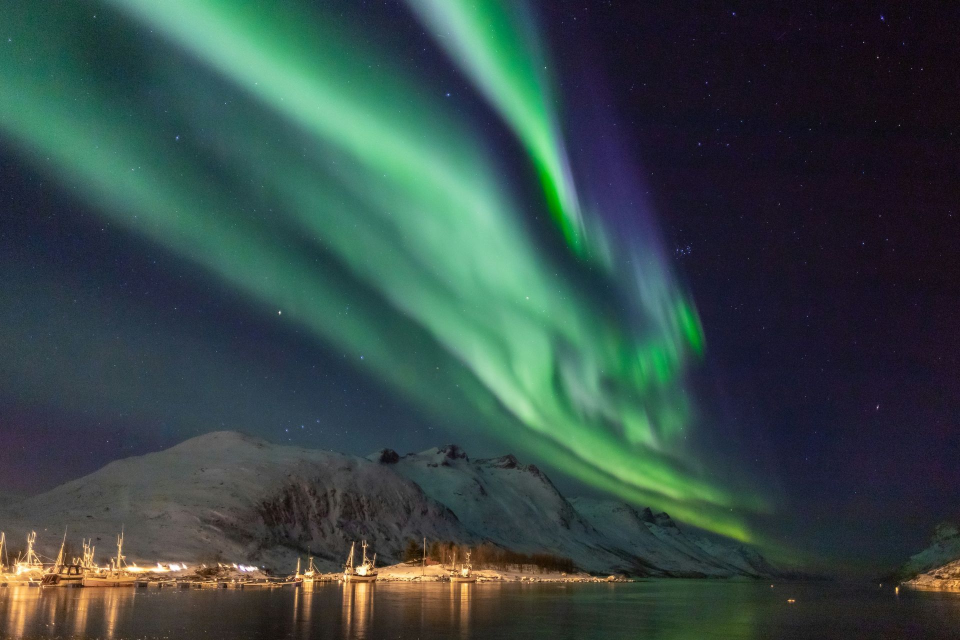 Green and blue aurora borealis lights over a snowy mountain range and still water over the city of Tromsø, Norway.