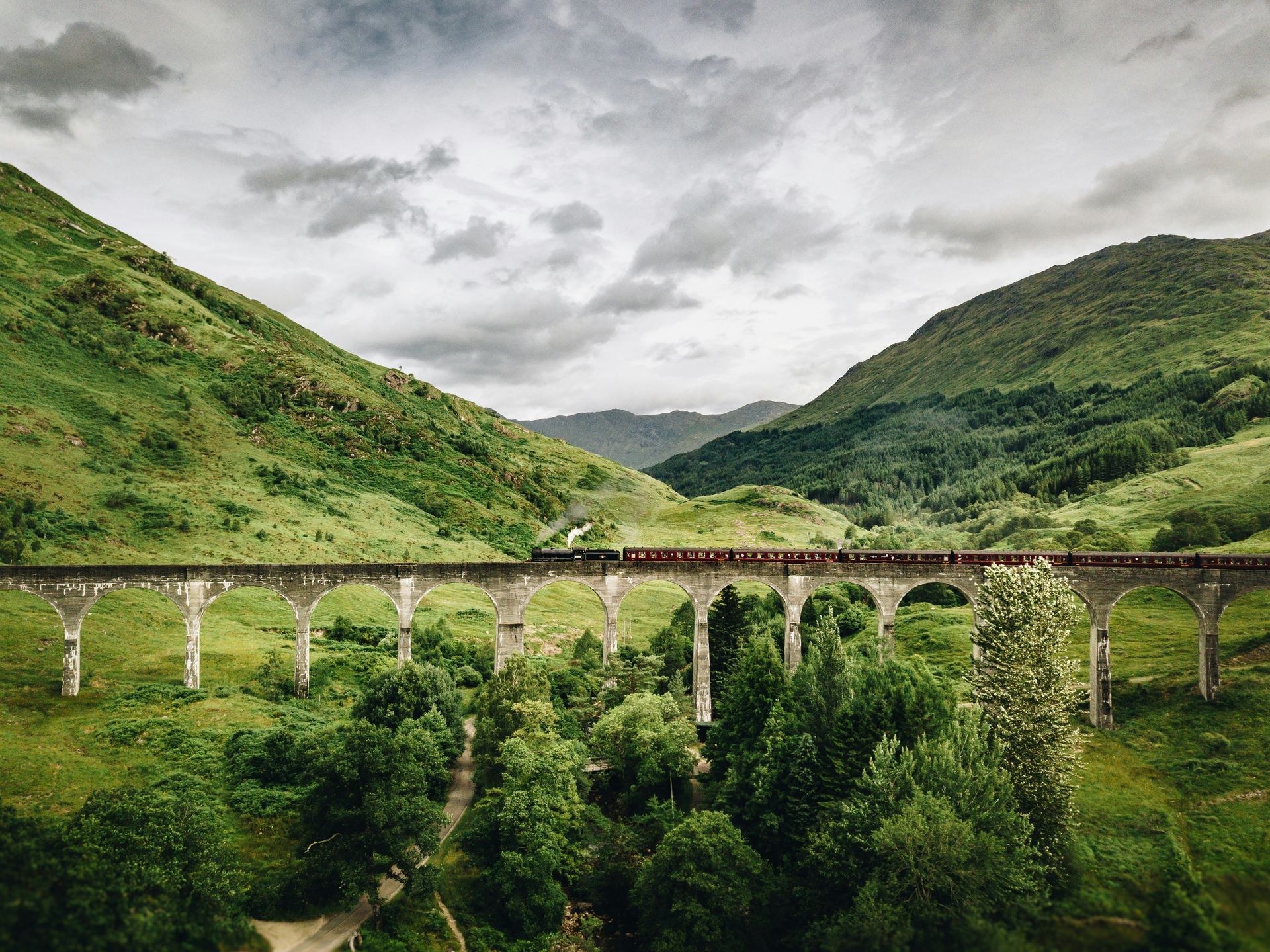 Train crossing the Glenfinnan Viaduct in Scotland, surrounded by green hills under a cloudy sky.