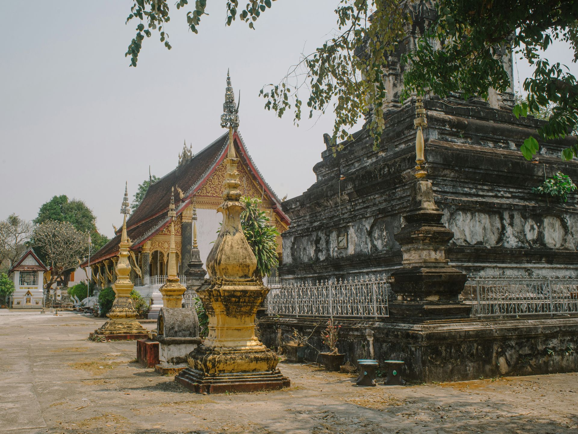 Temple complex with gold spires, ornate architecture, and a weathered, tiered structure in Luang Prabang, Laos.