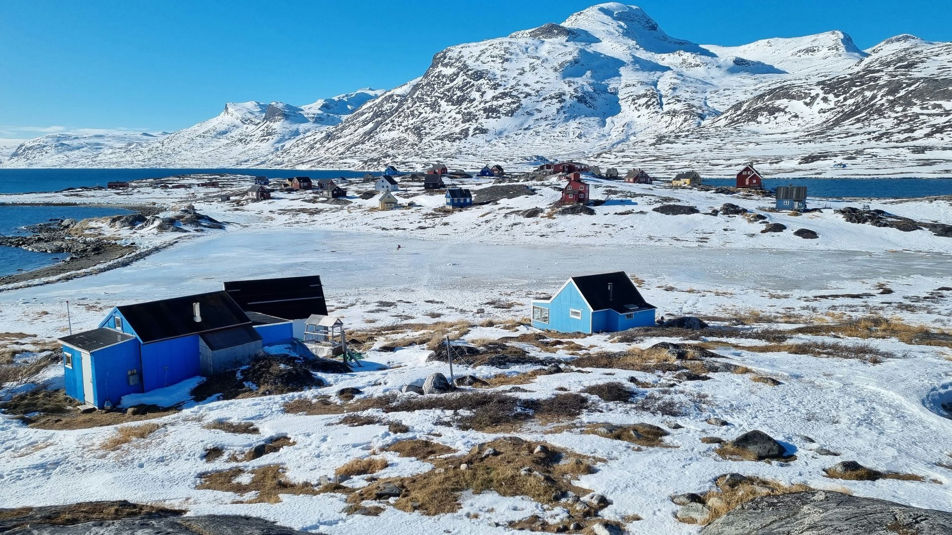 Snowy landscape with blue and brown huts near a coast, backed by mountains in Nuuk, Greenland.