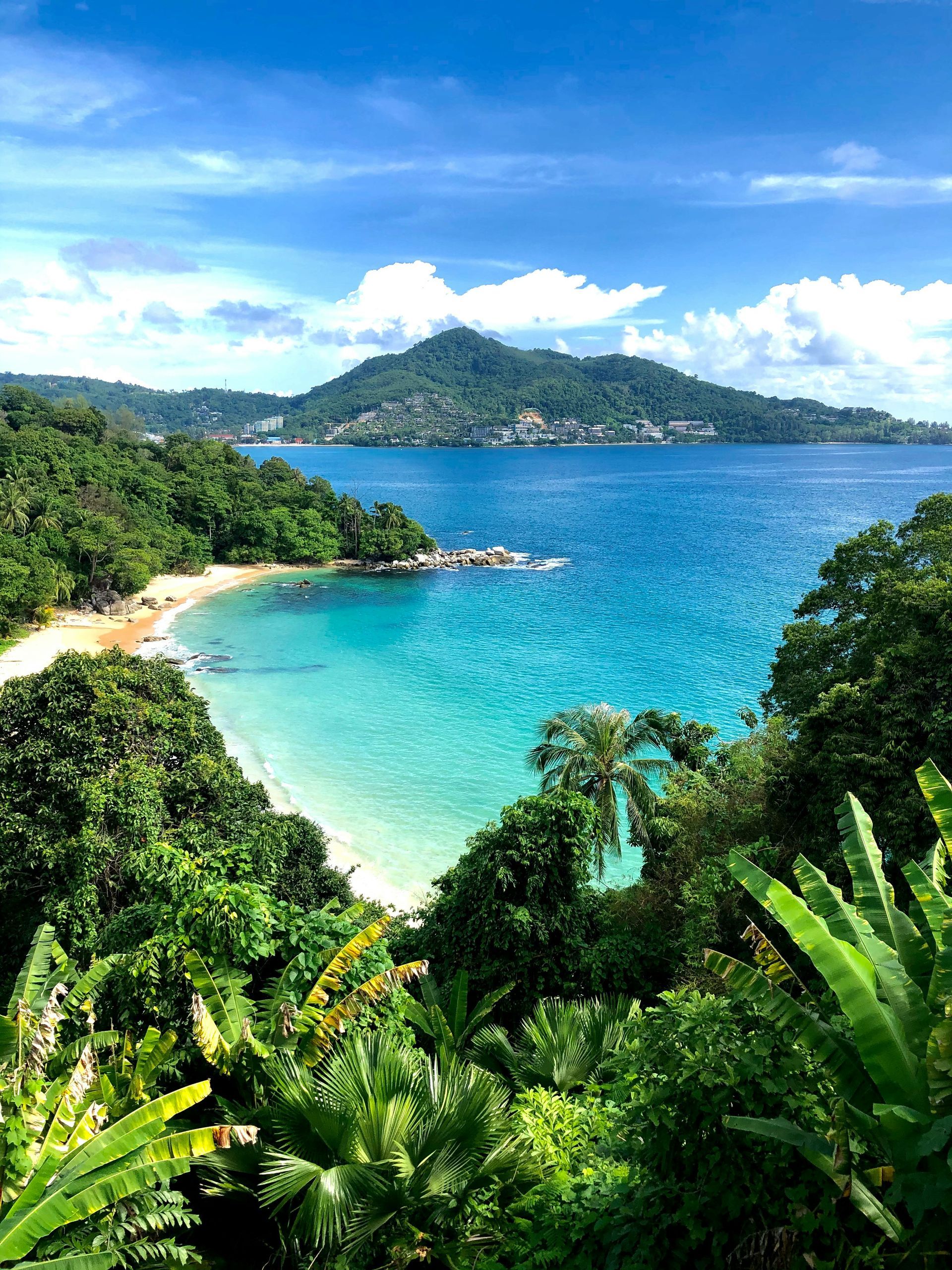 A view of a tropical beach with trees and mountains in the background in Phuket, Thailand.