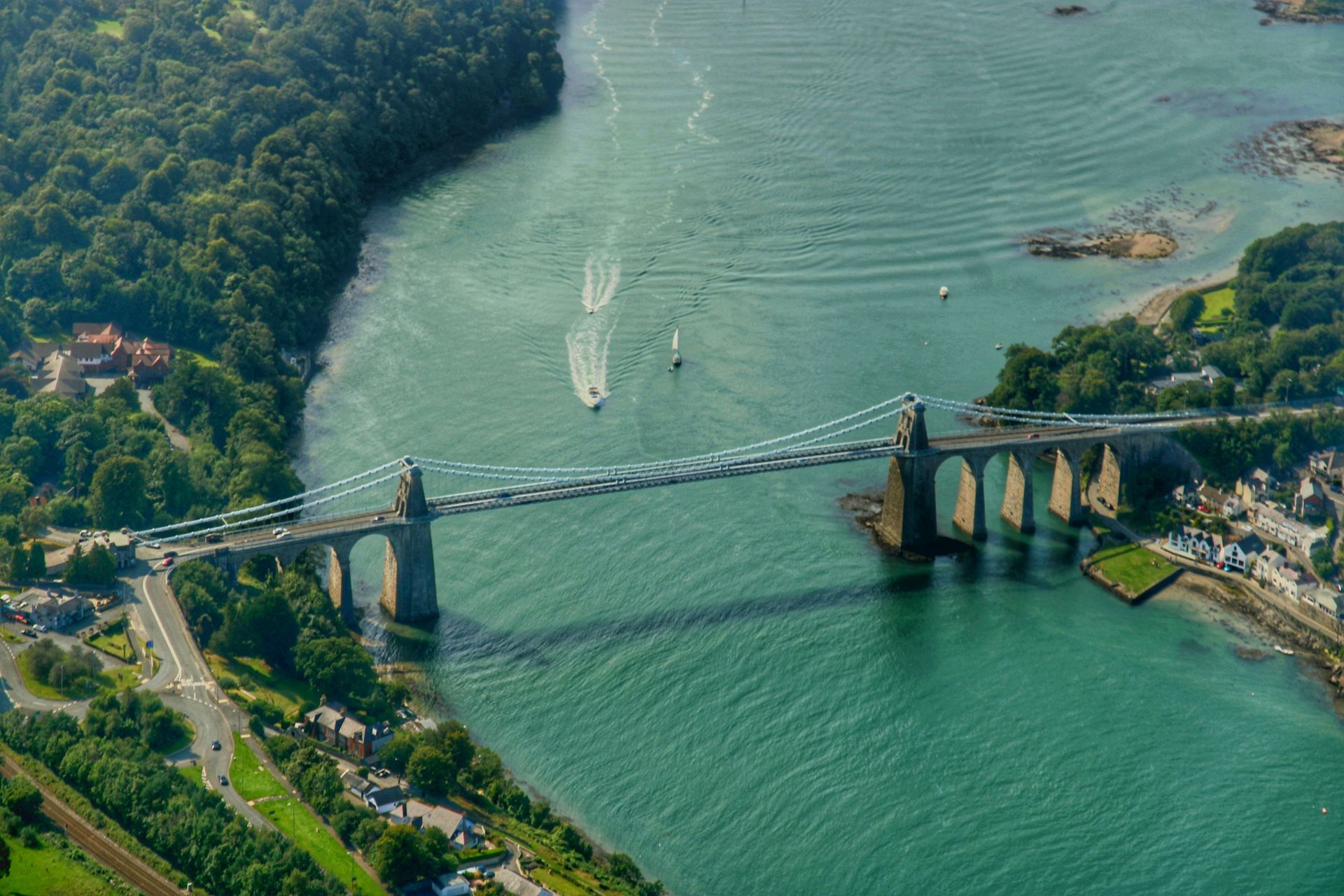 Aerial view of Menai Suspension Bridge in Wales spanning turquoise water, with boats and surrounding green landscape.
