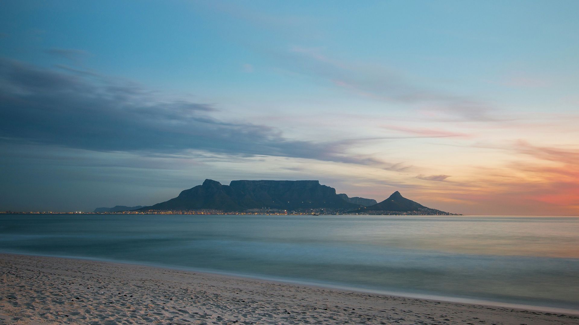 Table Mountain at sunset, viewed from a beach, with blue sea and orange sky in Cape Town, South Africa.