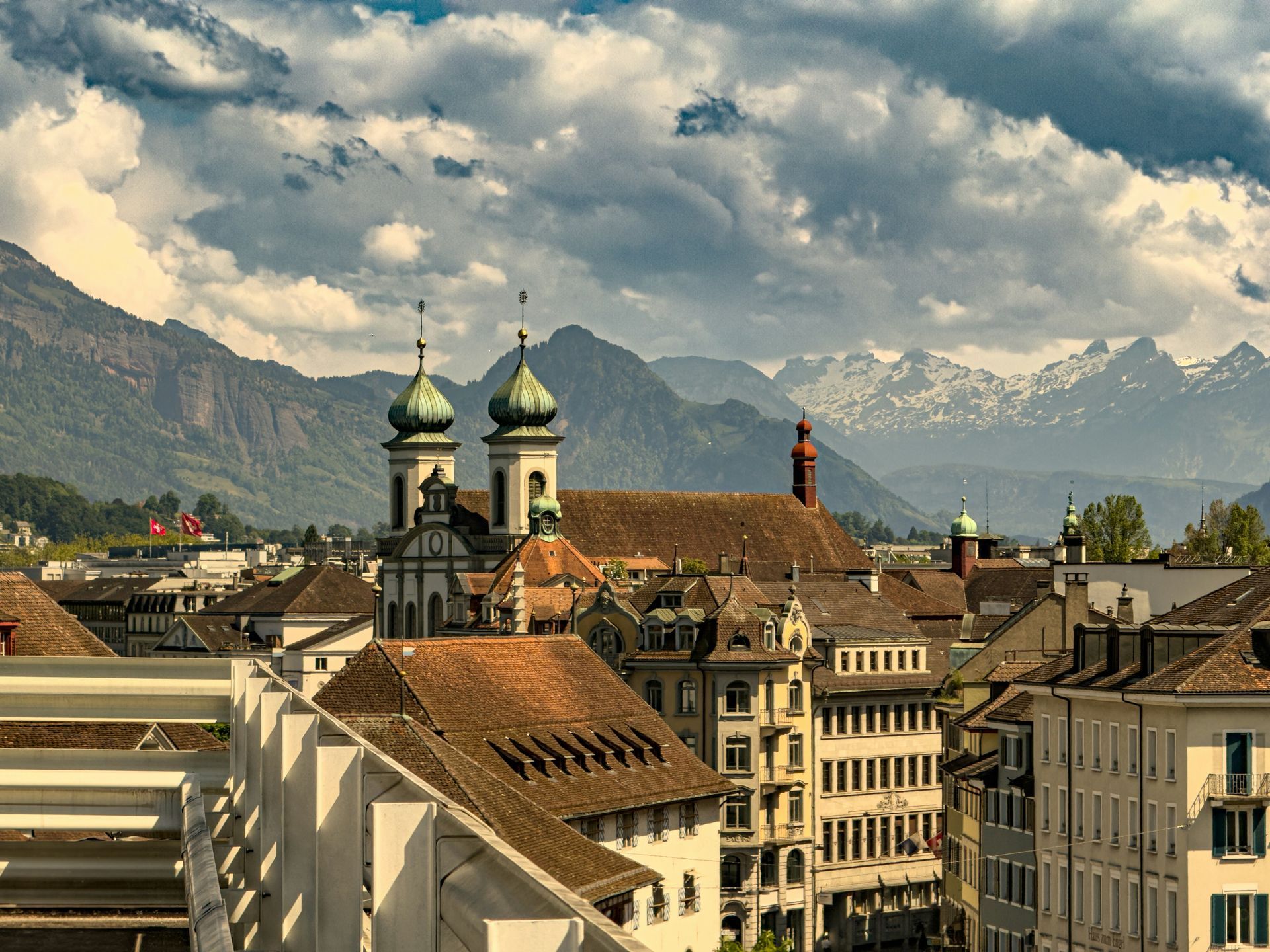 Cityscape with church spires, red rooftops, mountains in the background, and cloudy sky in Lucerne, Switzerland.