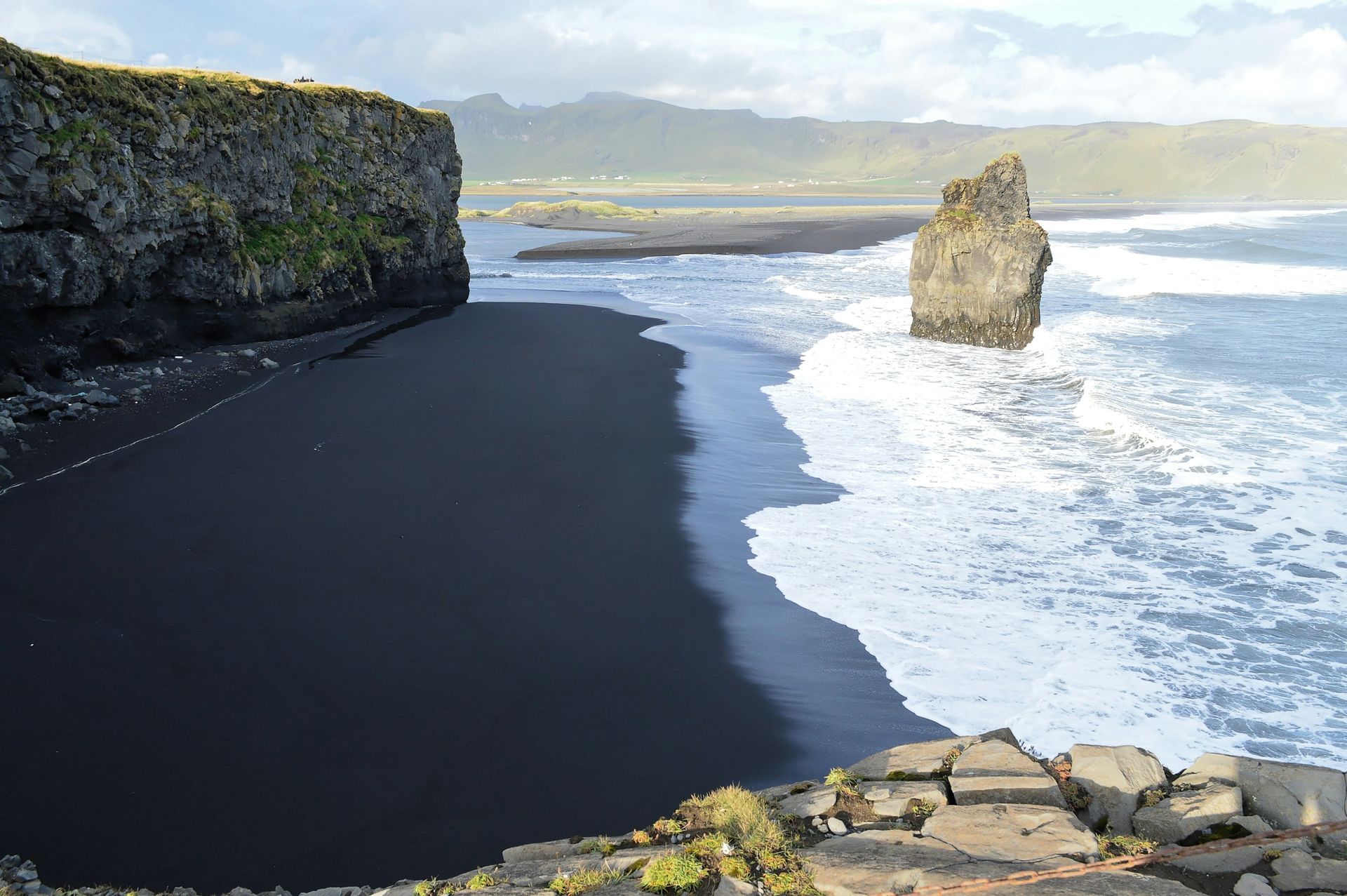 Black sand beach, rocky cliffs, ocean waves, and a large rock formation in Iceland.