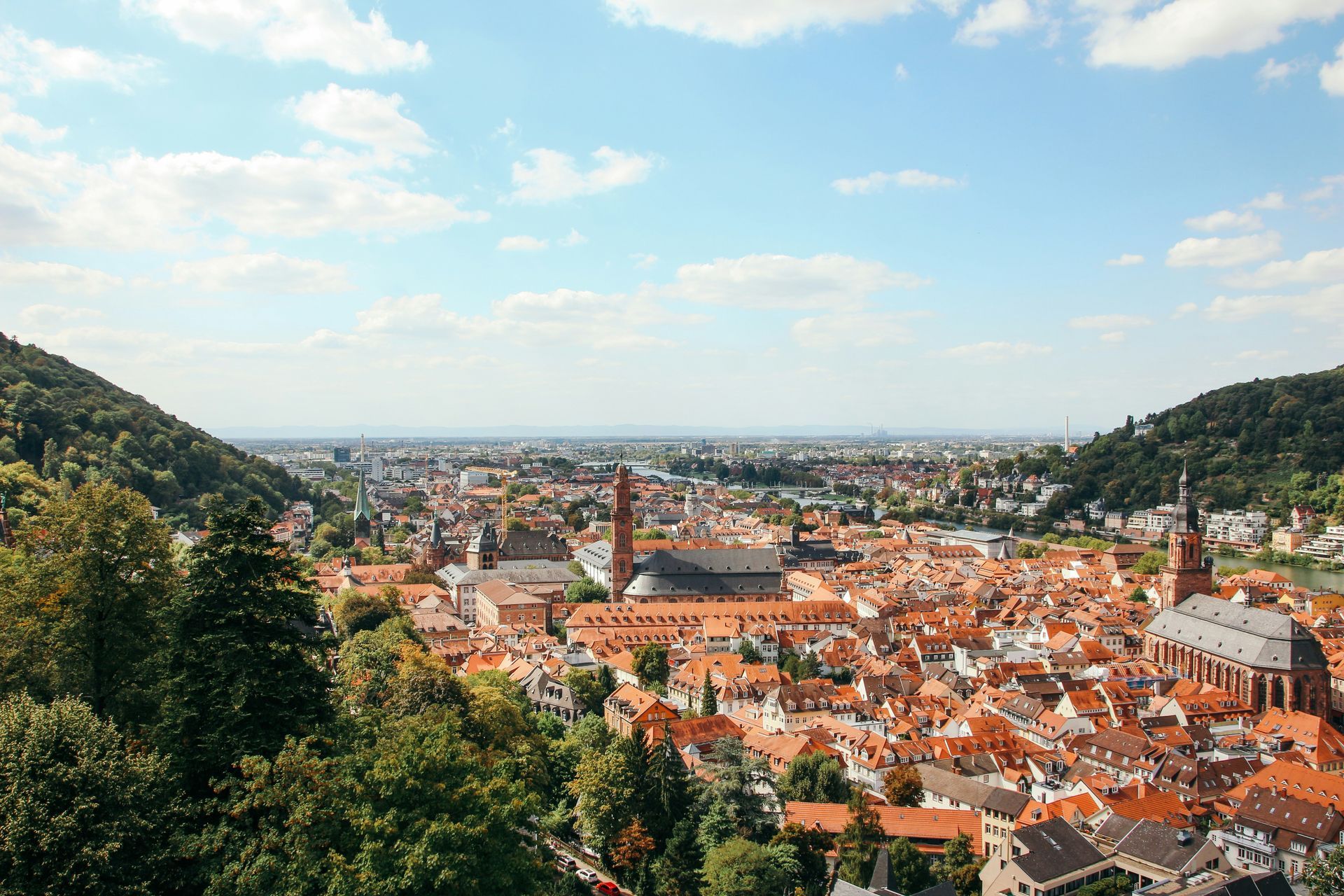 Panoramic view of Heidelberg, Germany, with red rooftops, a river, and hills under a blue sky.
